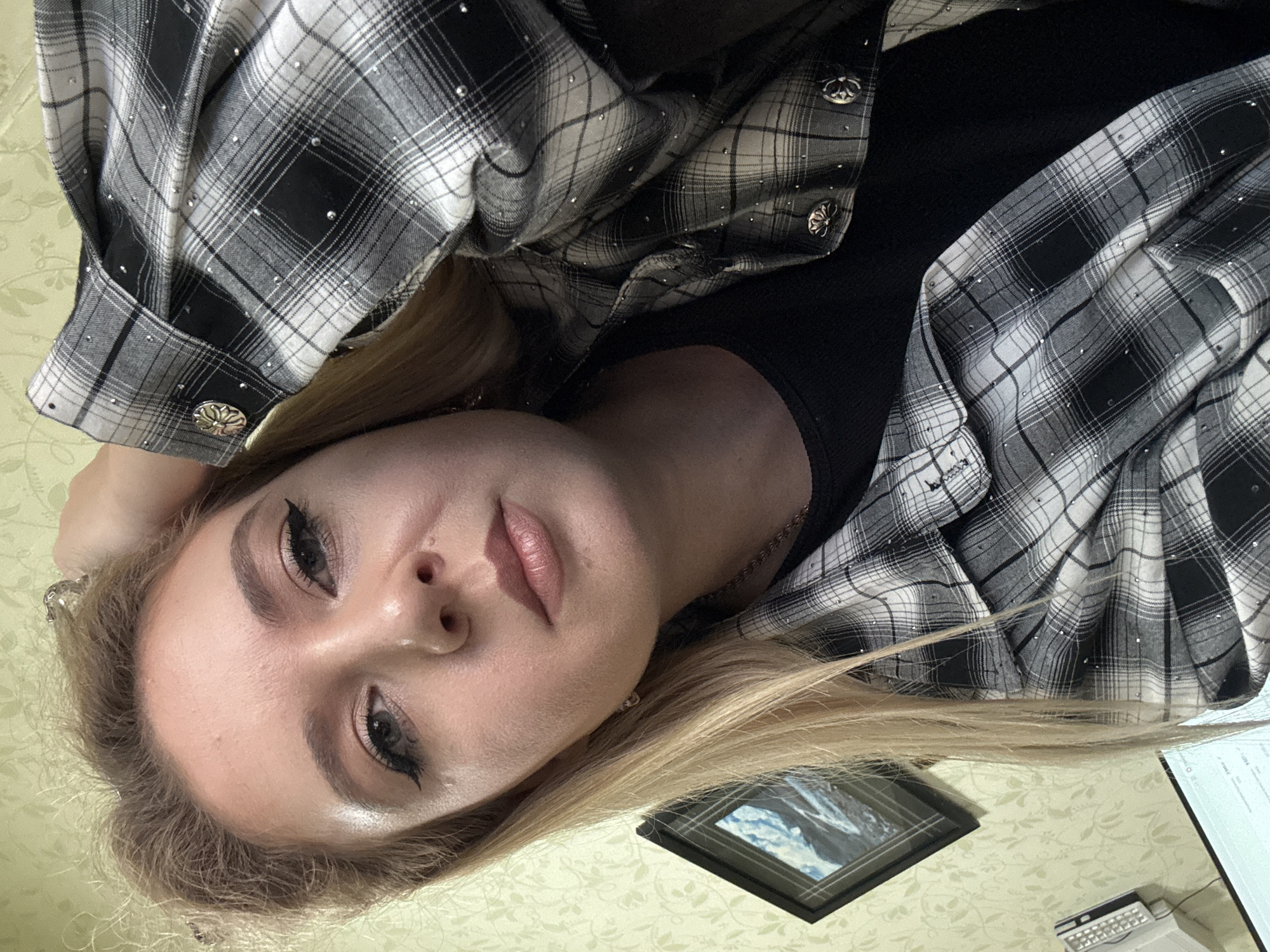 A young woman with blonde hair, wearing a black and white plaid shirt with rhinestones and a black undershirt, poses for a selfie indoors. Her right hand is resting on her head, and she has on dramatic winged eyeliner and lip gloss. The background features a wall with a floral pattern, a framed picture of mountains, and what appears to be a desk with a computer monitor and office equipment. The lighting is artificial and appears to be from overhead. The overall scene suggests a casual indoor setting, possibly an office or home environment.