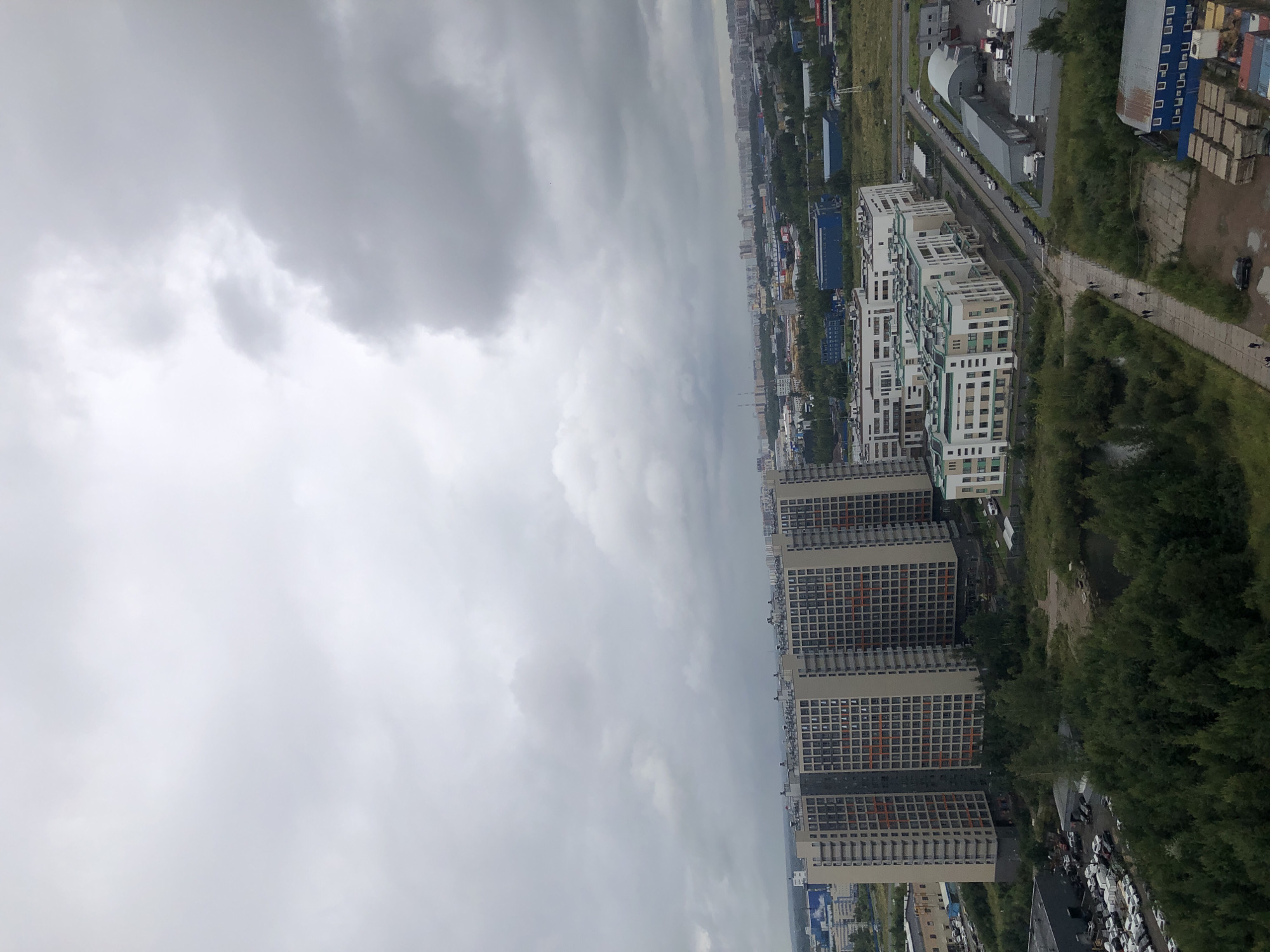An aerial, vertical shot shows a cityscape under a cloudy sky. The foreground is dominated by a dense cluster of green trees bordering a dark, still body of water. To the left of the trees, a large, gray, industrial-looking building is visible, with a parking lot filled with many white and dark-colored cars.
In the midground, a complex of tall, light-colored residential buildings with a grid-like pattern of windows and orange accents stands prominently. Adjacent to these buildings, a modern, low-rise complex of white and green buildings with numerous windows is situated. A road runs alongside this complex, lined with parked cars. Further back, a collection of industrial buildings, including some with curved roofs, can be seen.
The background reveals a sprawling urban landscape with many buildings stretching towards the horizon. The sky is filled with heavy, gray clouds, creating a diffused, overcast lighting condition. The scene appears to be a mix of residential and industrial areas within a city.