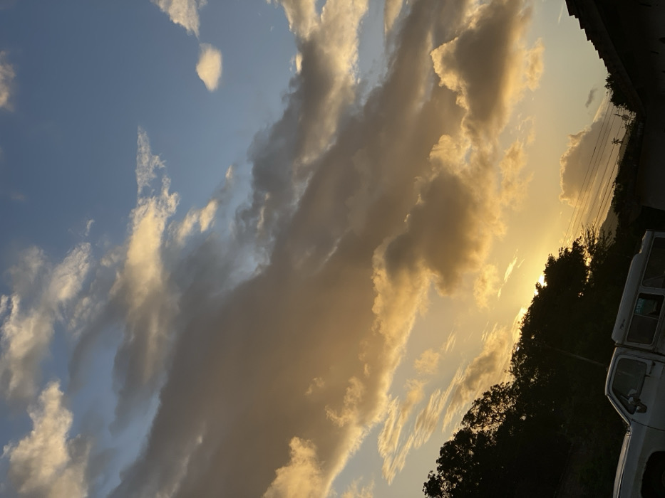 A landscape with a dramatic sky, featuring blue patches and layered clouds illuminated by the setting sun. The clouds range from wispy to thick and shadowed, tinged with golden hues. A line of trees and distant mountains silhouette against the bright light. A glimpse of utility poles and a white vehicle's side hint at civilization. The composition is rotated.