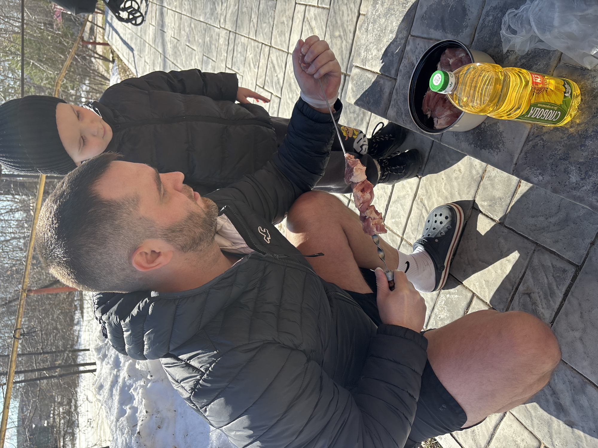 A man and a child are outside, likely preparing food on a patio or in a yard. The man, wearing a black puffer jacket and dark shorts, is seated on a grey brick surface, holding a metal skewer with pieces of meat on it. He wears white socks and dark grey Crocs. A young boy, wearing a black beanie and black puffer jacket, stands next to the man, looking at the skewer. A metal pot with meat inside sits next to the man, along with a bottle of cooking oil labeled "Слобода" (Sloboda). Patches of snow are visible on the ground, indicating it is winter. The background includes a wooden fence and bare trees. Based on the Cyrillic labeling of the cooking oil as "Слобода," which is a brand produced in Russia, the location is presumed to be in Russia, with the city of Chaikovsky listed as the location.