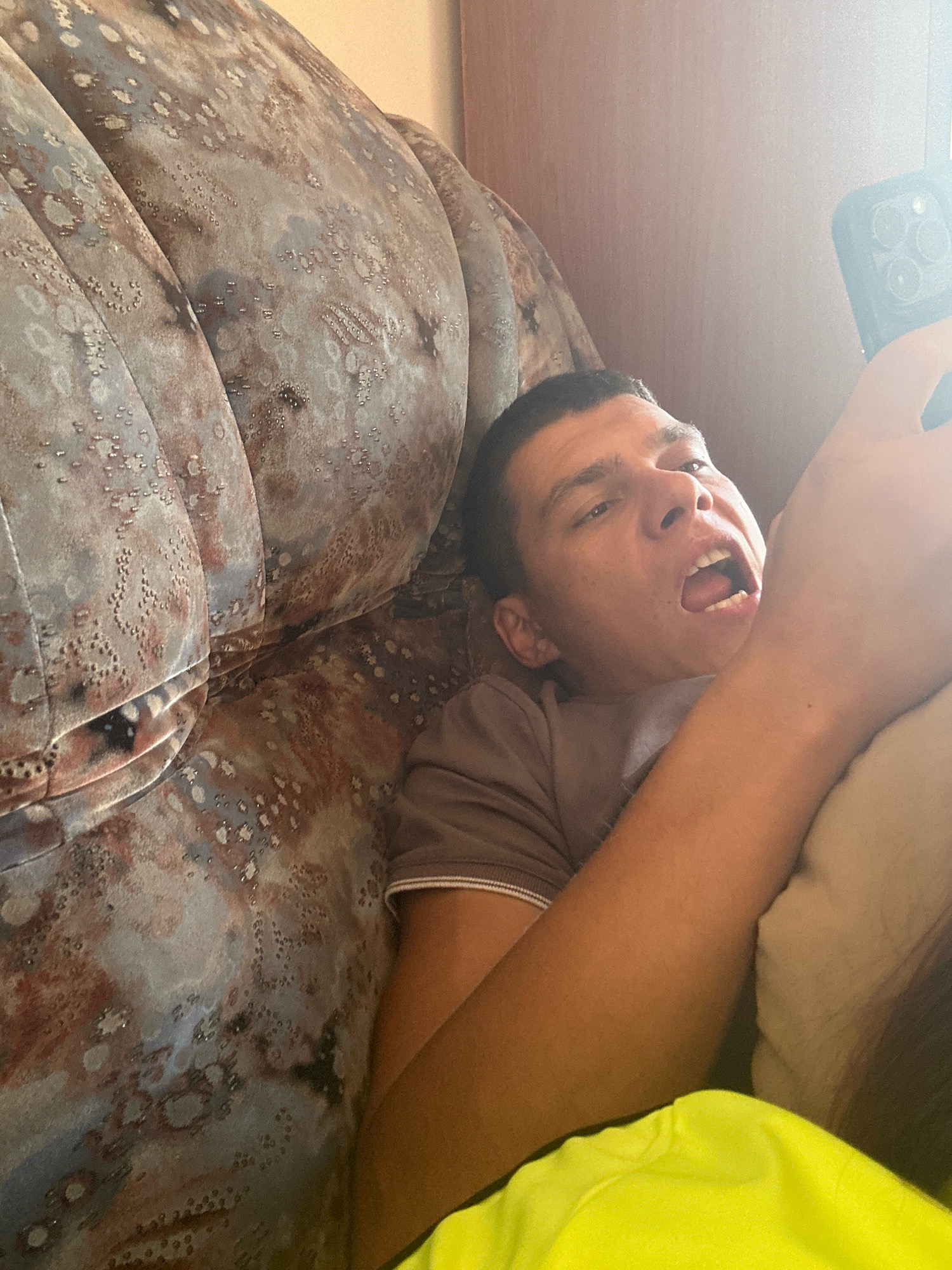 A young man is lying down on a patterned sofa, holding a smartphone in his right hand. He is looking at the phone with his mouth open. The sofa has a distressed, textured pattern with shades of brown, grey, and white. The man is wearing a brown t-shirt. A bright yellow garment is visible in the foreground. The lighting suggests an indoor setting with natural light entering from the right. The image does not contain information about the city of Gyumri, Armenia.