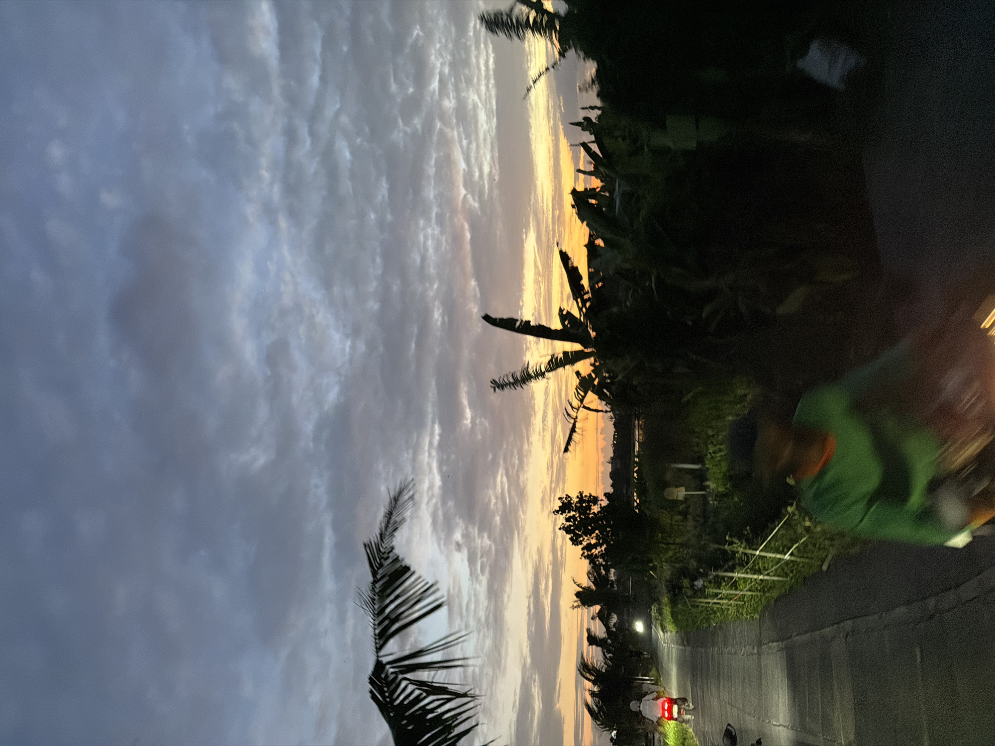 Captured at dusk in North Kuta, Indonesia, the image presents a scene along a road lined with lush tropical vegetation. The sky exhibits a gradient from dark, cloudy skies on the left to a vibrant orange sunset on the right, creating a dramatic backdrop. Silhouetted against the sunset are various palm trees and other foliage.
Along the road, there are two motorcycles, one with a red taillight indicating active operation, and another with a person in a green shirt who is out of focus. The road is illuminated by headlights, suggesting the fading daylight. The scene blends natural elements like the sunset and tropical plants with the man-made road and vehicles.