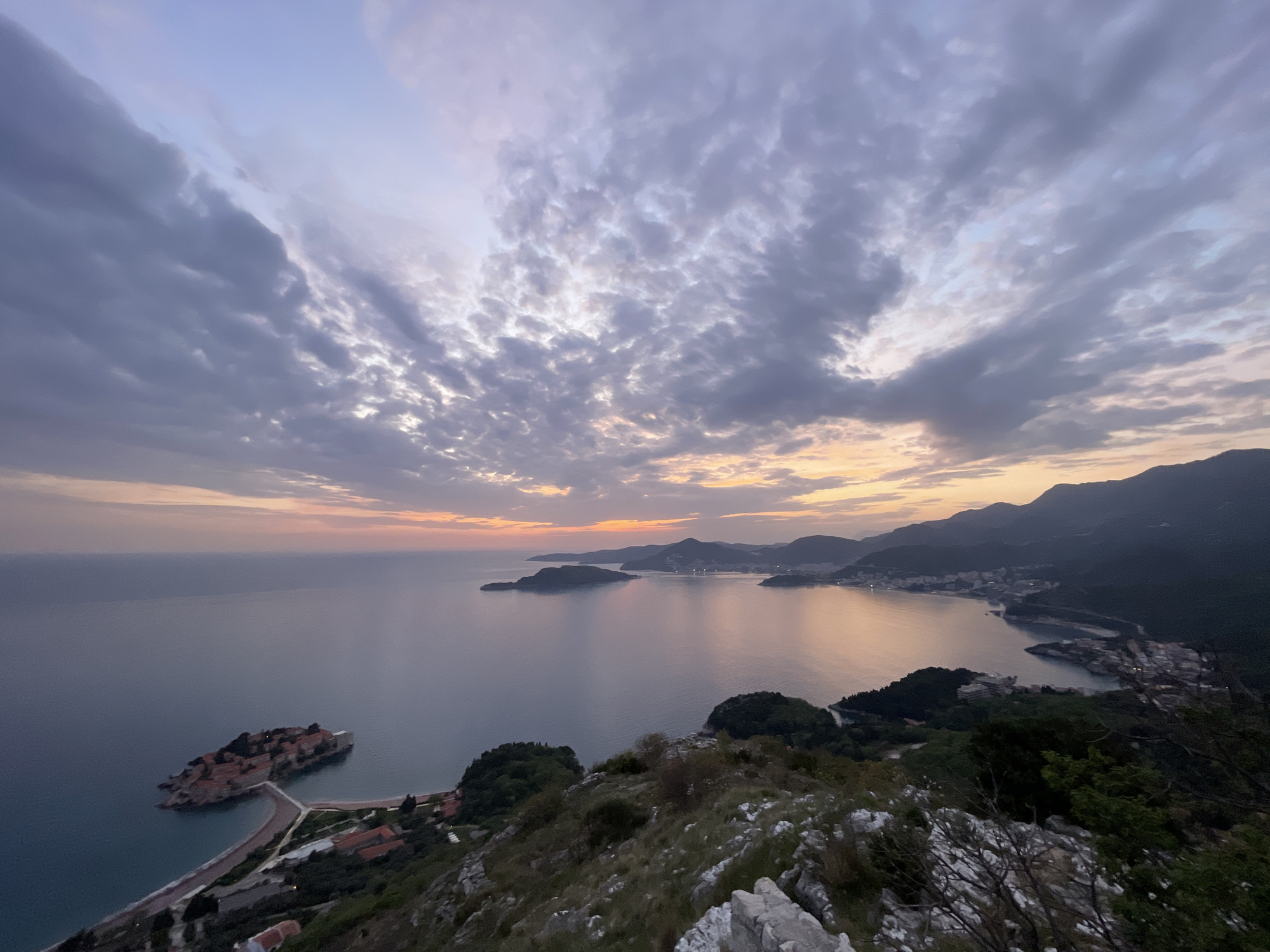 This expansive, elevated panoramic view captures the iconic Sveti Stefan island and the Montenegrin coastline, likely at sunset or sunrise. The foreground features rugged, rocky terrain with sparse vegetation, indicating a high vantage point.
The calm, reflective waters of the Adriatic Sea dominate the midground, extending towards the horizon. To the lower left, the distinct fortified island of Sveti Stefan is prominently featured, characterized by its clustered red-roofed buildings and connected to the mainland by a narrow causeway. Along the adjacent coastline, a beach and other coastal structures are visible.
Following the curve of the coastline to the right, several bays and coastal towns, some with visible lights, are nestled at the base of dark, undulating mountain ranges that recede into the distance. The sky above is a dramatic spectacle of clouds, ranging from dense, dark grey formations to lighter, broken patches. Warm pastel hues of orange, pink, and purple peek through near the horizon, signifying the early morning or late evening light. The overall scene conveys a sense of serene natural beauty and tranquility. No discernible text is visible in the image.
