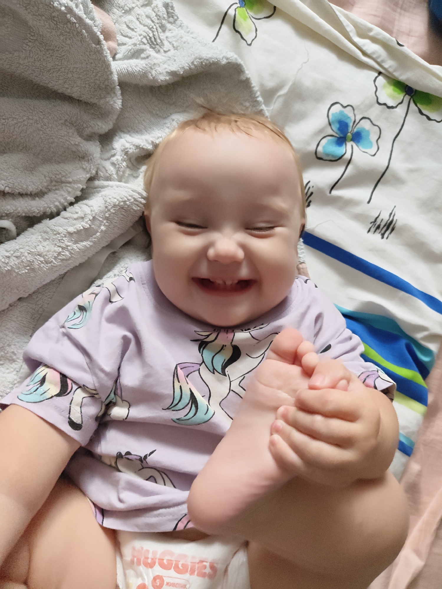 A close-up shot captures a happy baby lying on a bed. The baby is wearing a light purple t-shirt adorned with unicorns and a diaper. The baby's right foot is held by their hand, and their eyes are closed as they smile. The background consists of a light-colored blanket with a floral pattern and a textured grey blanket. The scene is indoors, likely in a bedroom in Krasnoobsk, Russia.