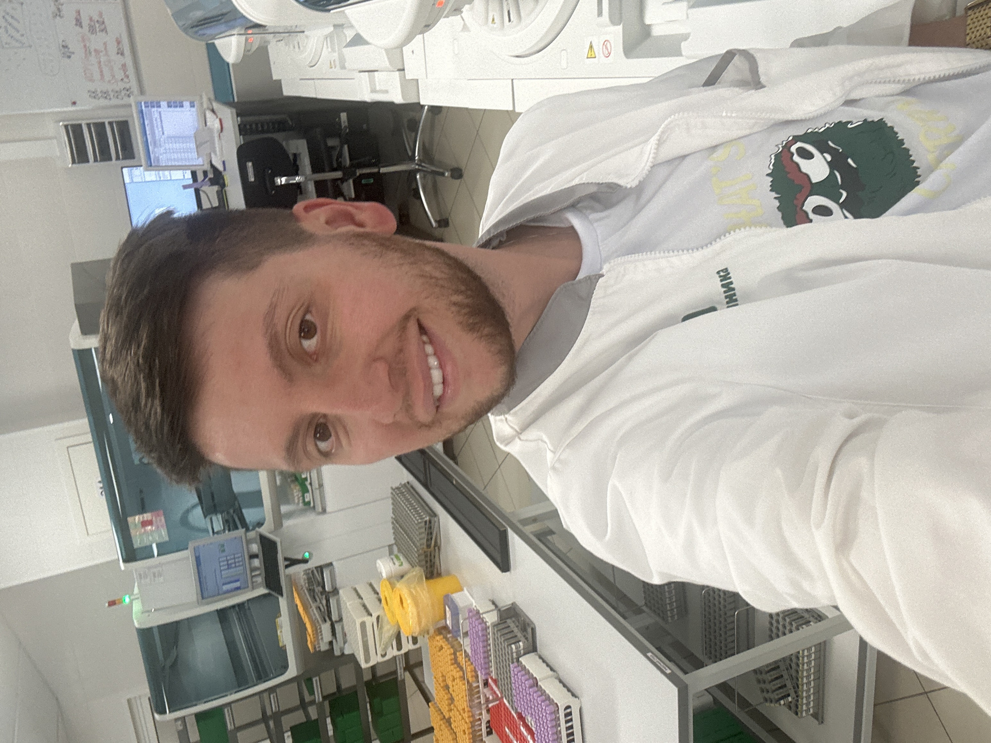 A young adult male, estimated to be in his late 20s to early 30s, with dark short hair, light brown eyes, and a short beard, is depicted smiling directly at the viewer. He is wearing a white zip-up lab jacket over a white t-shirt, which features a graphic of a green, furry cartoon monster and partial yellow text.
The scene is a clinical or research laboratory environment, situated in Moscow, Russia. The background includes advanced laboratory automation systems; to the left, a large analyzer with a display screen is visible, accompanied by racks containing numerous test tubes with yellow, purple, and red caps, yellow biohazard waste bins, and metal storage racks on a white countertop. Green shelves are positioned further back. To the right, additional laboratory equipment is present, along with a whiteboard containing handwritten notes and two computer monitors on a desk displaying digital information. The area is well-lit, characterized by white walls and light-colored floor tiling, indicative of a professional and sterile work setting.