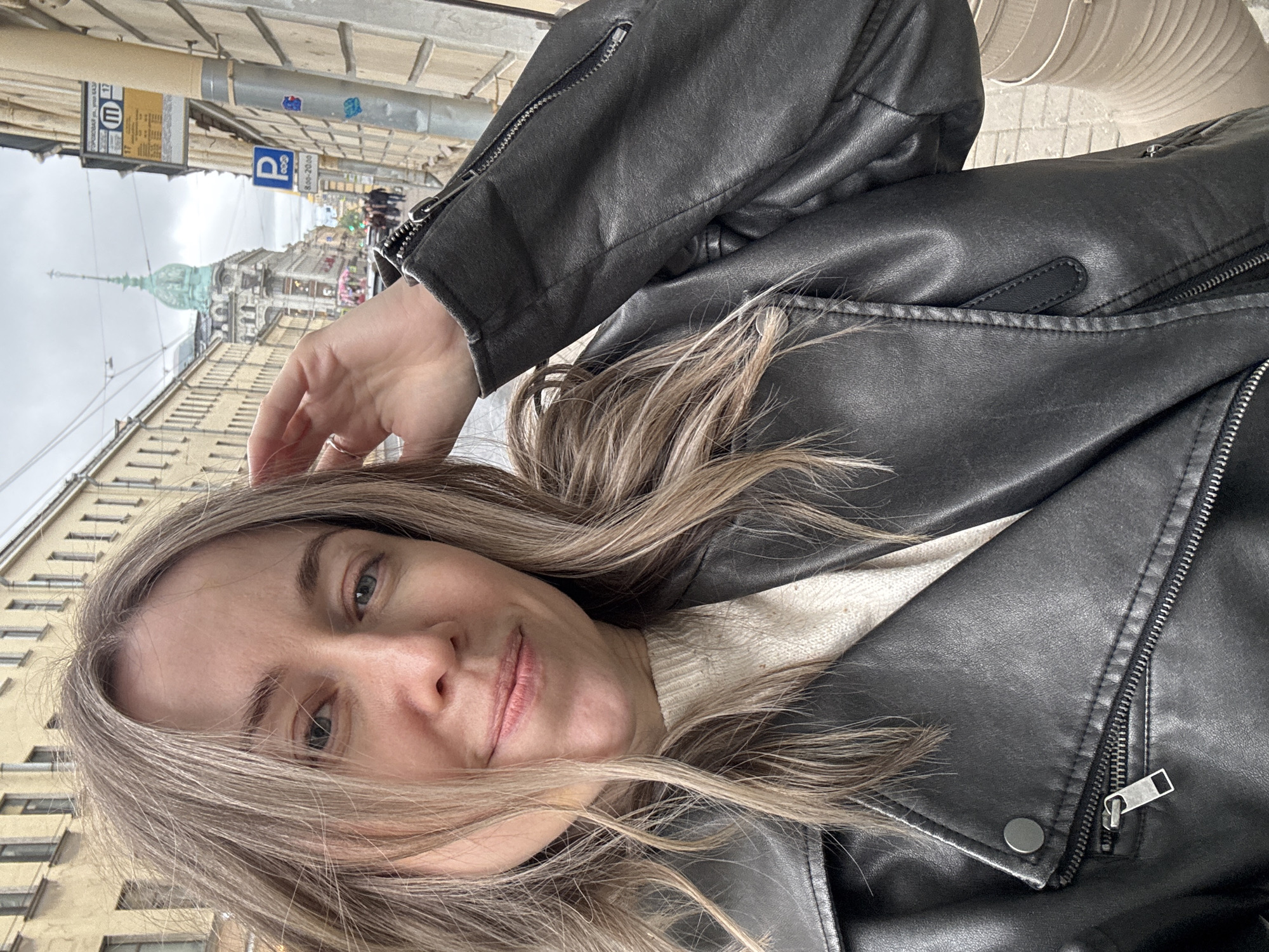 A selfie-style photograph captures a person against the backdrop of a Saint Petersburg street. The individual, wearing a black leather jacket over a light-colored sweater, has blonde hair and is looking towards the camera. The background shows a street with buildings characteristic of Saint Petersburg's architecture, including a building with a distinctive dome. Street signs and pedestrians are visible in the distance. The weather appears overcast.