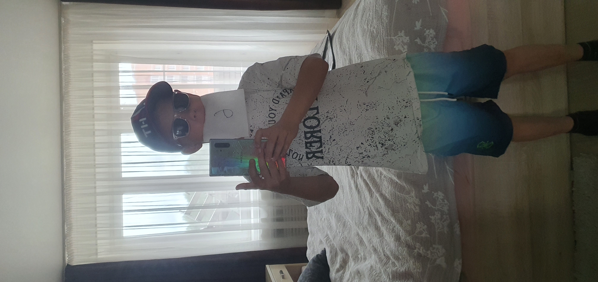 A person wearing a cap, sunglasses, a white t-shirt, and blue shorts is taking a selfie with a smartphone. The person is standing in a room with white walls and a window with blinds. A bed with white bedding is visible in the background. The image was taken in Chaikovsky, Russia.