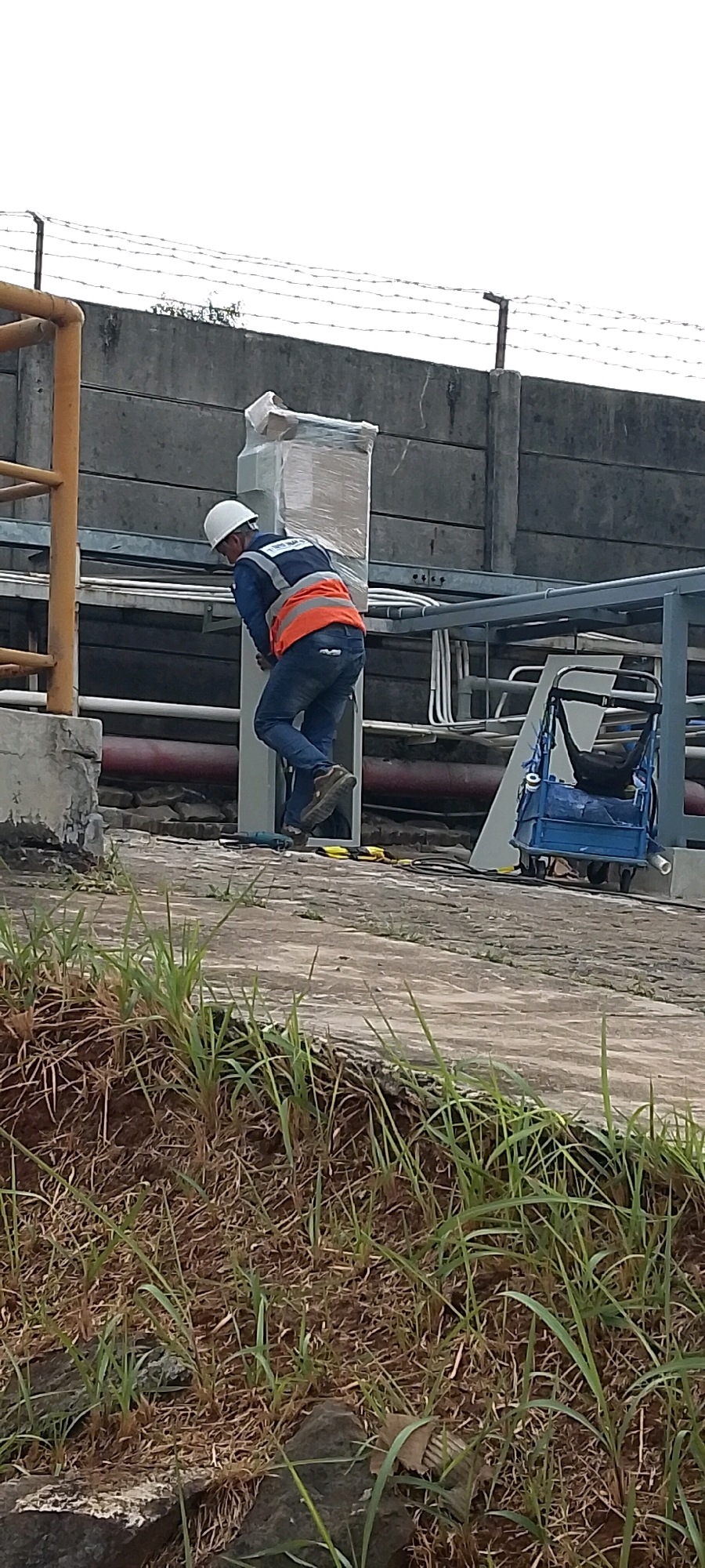 A lone worker, wearing a white hard hat and a blue shirt with an orange safety vest, is shown from the rear as he works on a vertical metal structure. The worker is leaning into the structure, with his right leg bent and his left leg extended, appearing to be in a precarious position. The structure he is working on is encased in plastic wrap and appears to be a utility box or junction. The background consists of a concrete wall topped with barbed wire, suggesting an industrial or secure area. Several pipes and conduits are visible running along the wall and overhead. To the right of the worker, a blue industrial cart with a black bag on it is parked. The ground in the foreground is a concrete walkway with some overgrown grass and rocks along the edge. The scene is set outdoors, possibly in a manufacturing plant or industrial complex, within the city of Depok, Indonesia.