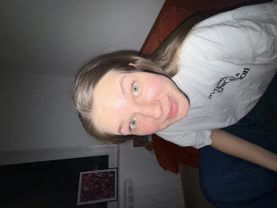 A woman with light brown hair and green eyes smiles at the camera. She wears a white t-shirt with "It's Time for a Vacation" printed in black. The background includes a dark gray wall, a window with a white frame, and a framed piece of art with red and white abstract patterns. A red couch is partially visible behind her, and she seems to be sitting on a dark blue surface. The lighting is dim, creating shadows in the room.
