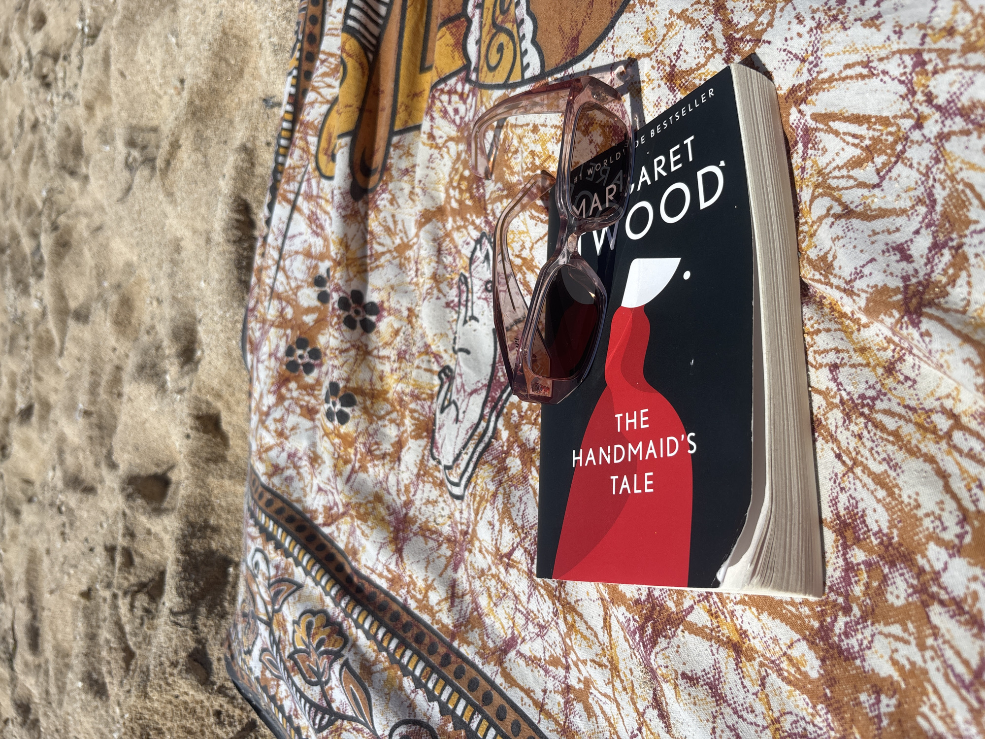 The image depicts a book titled "The Handmaid's Tale" by Margaret Atwood, with sunglasses resting on top of it. The book and sunglasses are placed on a patterned cloth or textile. The background includes sand, suggesting a beach setting. Based on the location provided, the scene is likely on a beach in Kuta Selatan, Indonesia. There are no people visible in the frame.