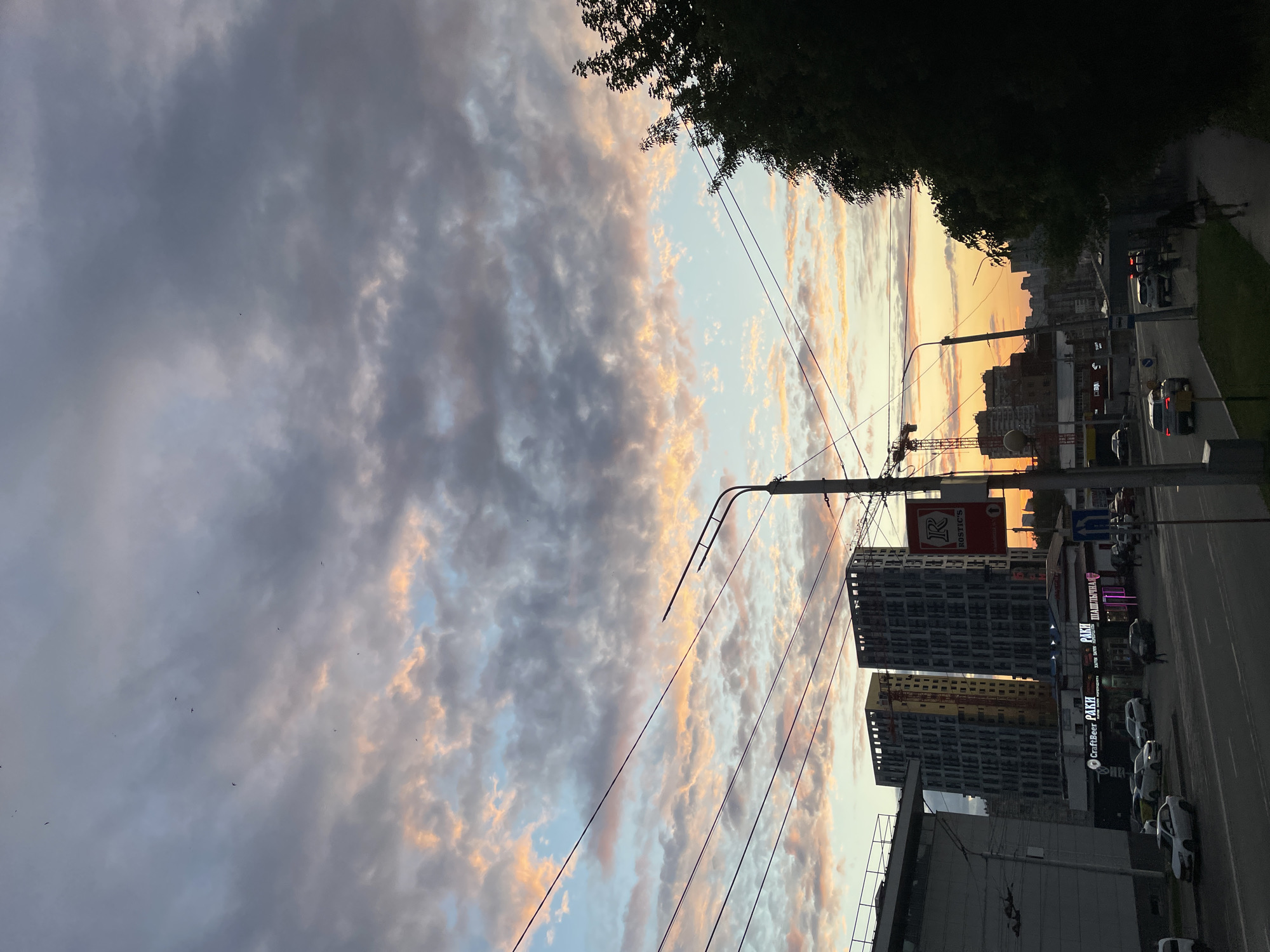 The image depicts an urban street scene in Ufa, Russia, at sunset. The sky is filled with dramatic clouds tinged with orange and pink hues, indicating the time of day. Power lines crisscross the frame, and a lamppost stands to the right. In the background, several multi-story buildings are visible, some appearing to be under construction with cranes present. A street with cars and a few pedestrians is in the foreground. Signs for businesses like "CraftBeer" and "RAKI" are partially legible on the lower buildings. The overall atmosphere suggests a typical evening in a Russian city.