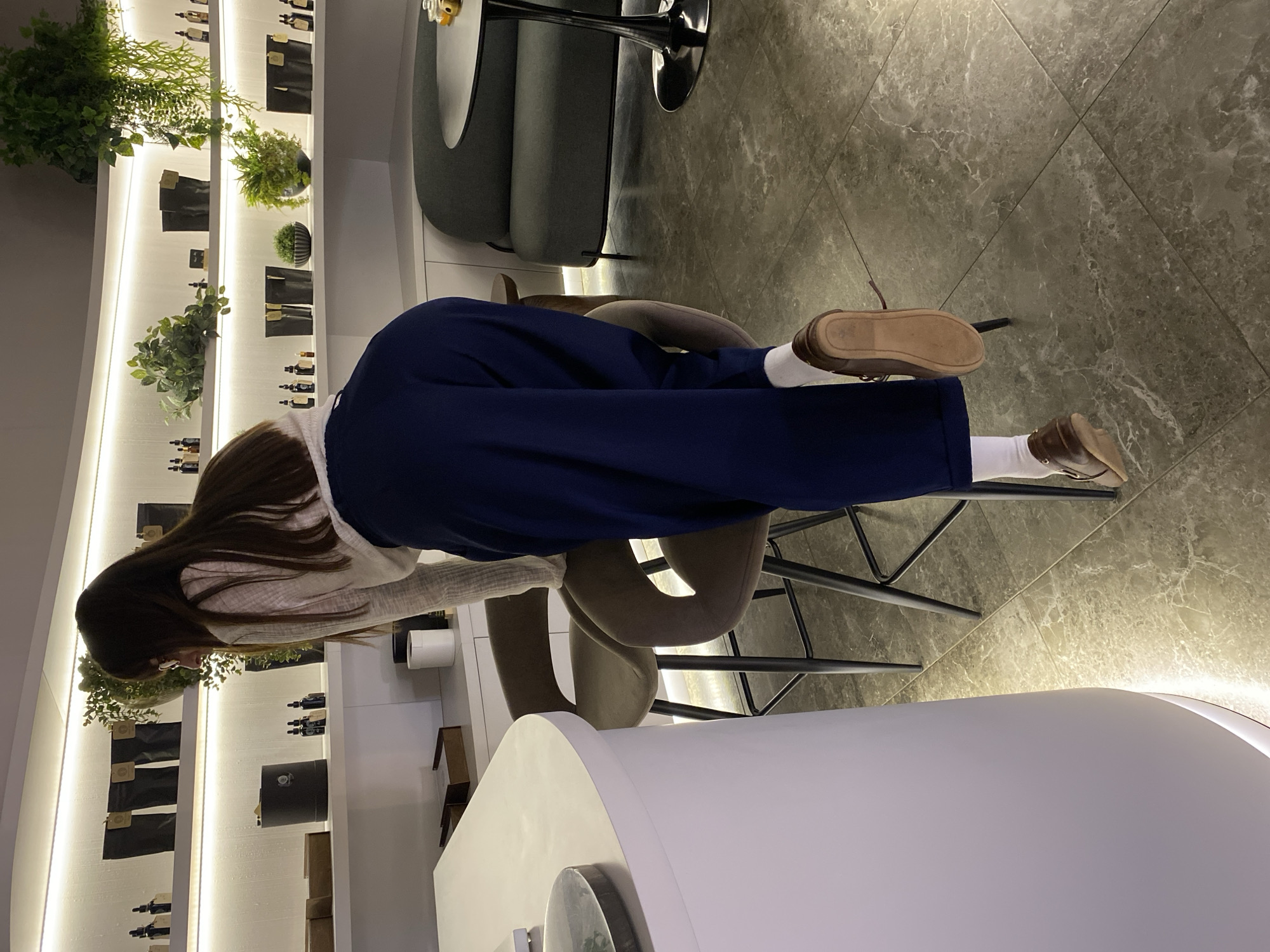 This media file captures an indoor scene within a modern retail space, possibly a boutique or a cosmetics/perfume store, located in Votkinsk, Russia. The central subject is a person, seen from behind, with long, dark hair, wearing a light-colored top, dark blue loose-fitting pants, white socks, and brown leather shoes. They are leaning over a white counter in the foreground, intently examining products displayed on a brightly illuminated shelving unit.
The shelving unit, mounted on a white wall, features multiple tiers with integrated warm LED strip lighting. Various small dark bottles, likely perfumes or essential oils, are neatly arranged on the shelves, some accompanied by small black pouches or packaging. Several small potted plants are interspersed among the products, adding a touch of greenery to the clean aesthetic.
The flooring consists of large grey tiles with a subtle white marble-like pattern. In the background, a dark grey upholstered lounge chair and a small round table are visible, suggesting a seating area within the store. Two dark-framed bar stools or high chairs are partially visible beneath the person, with one of the person's feet resting on a stool's footrest. The overall setting appears modern, clean, and well-lit, creating an inviting atmosphere for browsing. No distinct text is clearly legible on any of the products or signage.