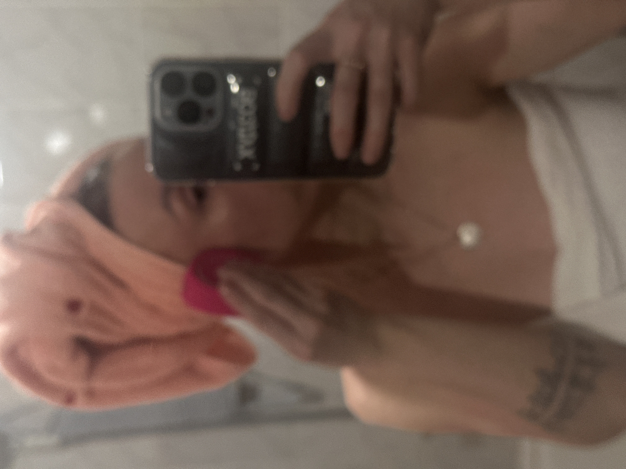 A person is captured in a mirror selfie, seen from the chest up, with the image rotated sideways. They are wearing a white towel wrapped around their body and a peach-colored towel on their head, suggesting they may have recently showered or are engaged in a self-care routine. The individual is holding a black smartphone with a triple-lens camera system, likely an iPhone, in their right hand, using it to take the photo. A ring is visible on a finger of the hand holding the phone. With their left hand, the person is using a vibrant pink beauty sponge or applicator to apply a product to their face. A silver-colored necklace with a circular pendant hangs around their neck. A tattoo is visible on their left arm/shoulder area. The back of the phone case features stylized text that appears to read "CREATOR." The background is softly blurred, showing what appears to be an indoor setting, likely a bathroom, given the towels and mirror. The overall lighting is subdued.