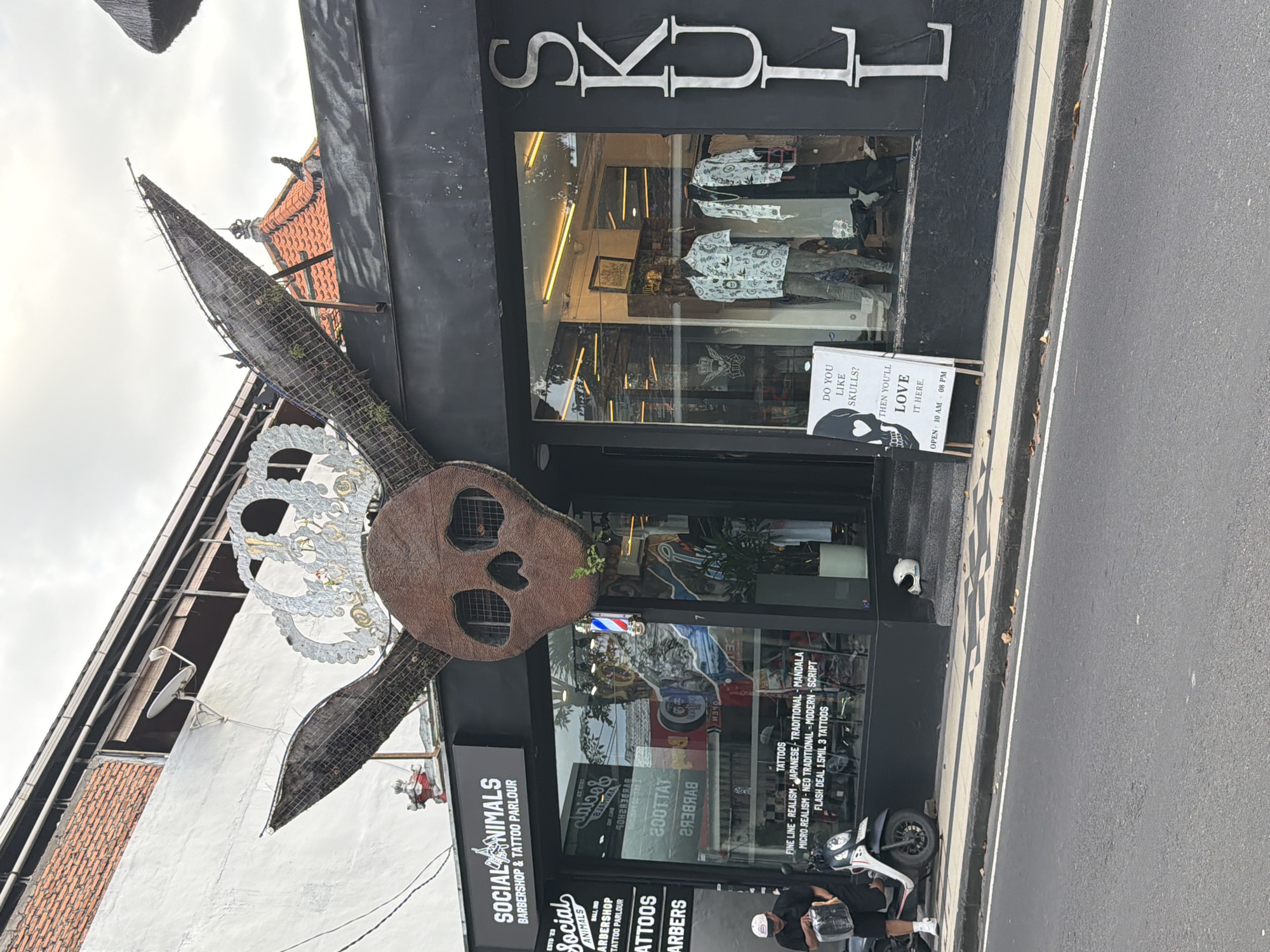 The image shows the exterior of a business called "Social Animals Barbershop & Tattoo Parlour" in Kuta, Indonesia. Above the entrance is a large, rusted metal skull with rabbit ears. The business's name and services are displayed on a sign. There is a sign to the side of the door. A motorcycle is parked out front, and a person is sitting nearby. The building has a black facade with large windows displaying clothing and tattoo artwork. The name "SKULL" is vertically aligned above the shop. A barber pole is visible in the storefront.
