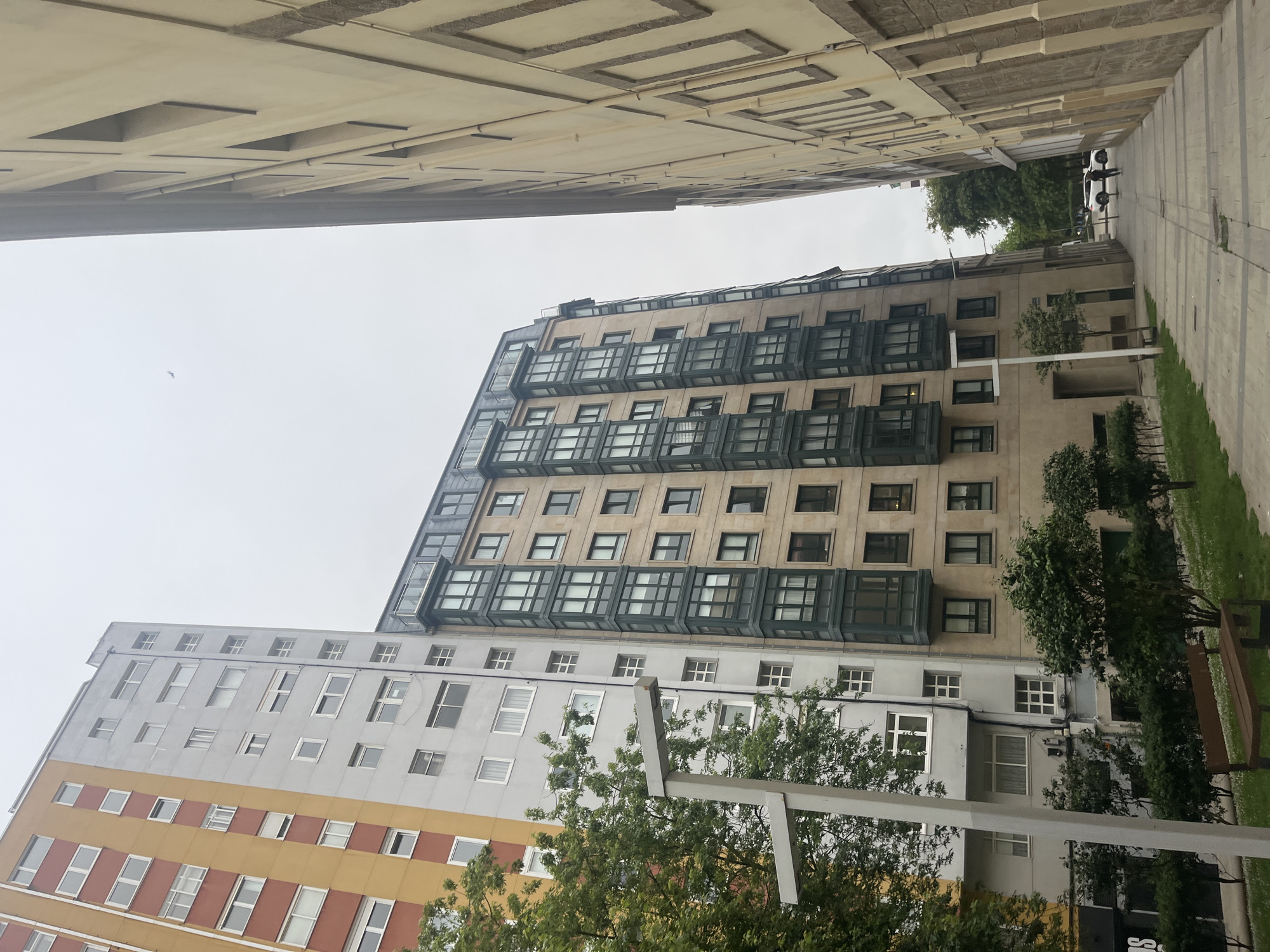 The image shows a street scene in A Coruña, Spain, with a view looking upwards between buildings. A multi-story beige building with green-framed windows and balconies is visible. To its left is a taller building painted white, gold, and rust with several windows. Part of another building is seen at the very top of the image, obscuring much of the sky. A paved sidewalk with a grassy area and a few benches lines the street. A modern streetlight or architectural element is in the foreground, partially obscured by trees. Two individuals are walking away from the camera in the distance. The weather appears overcast.