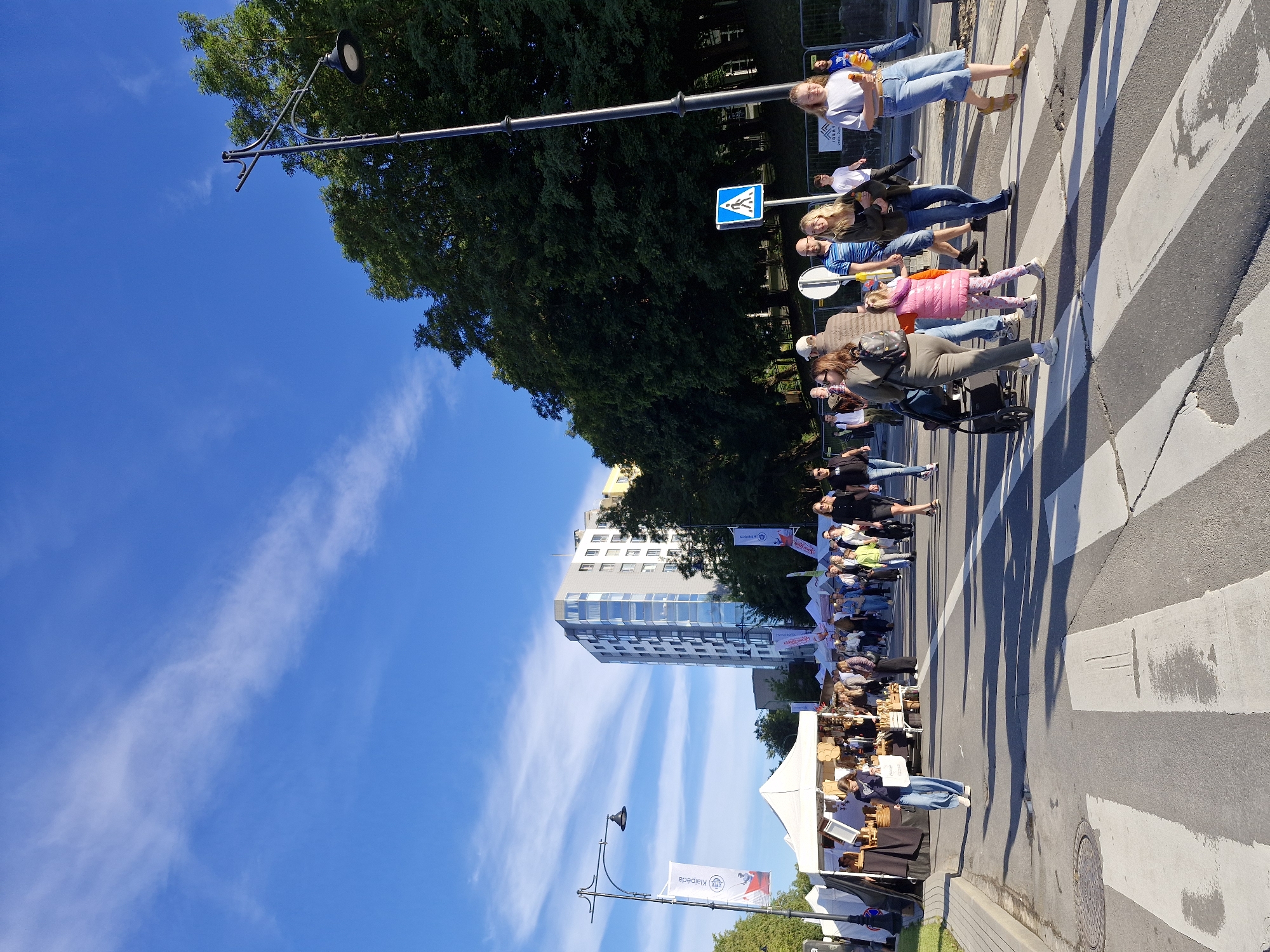 The image captures a street scene in Klaipėda, Lithuania, on a bright, sunny day with a few wispy clouds in a vibrant blue sky. The perspective is from a low angle, looking upwards towards a street lamp and a line of trees. In the foreground, a pedestrian crossing is clearly visible, with several people walking across it or standing nearby. The people are diverse in age and attire, suggesting a casual event or outing. Some individuals are holding items, and one person is pushing a stroller.
Behind the people and the crossing, a modern, multi-story building stands out against the trees. Further back, white tents suggest an outdoor market or event. A street lamp with a black fixture is prominent in the upper left quadrant of the image, its arm extending over the scene.
Several flags with text are visible, adding to the context of a public gathering. One flag, partially obscured, appears to have text that might relate to the location or event. The overall atmosphere seems relaxed and cheerful, with people enjoying the outdoor conditions. The presence of the pedestrian crossing, the building, and the flags with the specific text "Klaipėda" clearly indicate the location. The lighting suggests it is daytime, likely mid-morning to afternoon, given the strong shadows cast by the people and the brightness of the sky.