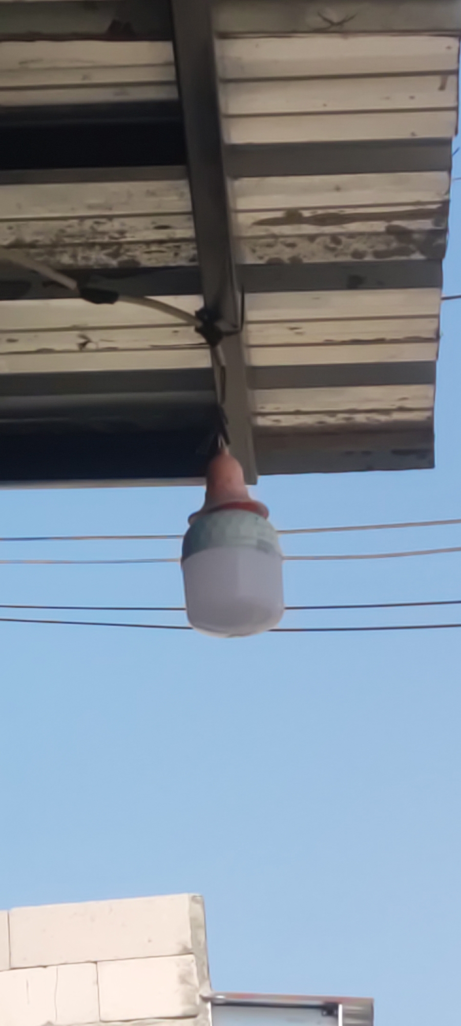 A low-angle shot captures a light fixture suspended from a corrugated metal awning, its white and light green casing a stark contrast against the clear blue sky. The light dangles precariously from wires that run horizontally across the frame. The awning, a weathered and slightly damaged structure, is attached to a metal frame. In the lower portion of the image, a section of a concrete block wall, typical of urban construction in Gebang, Indonesia, is visible under the blue sky. The overall scene depicts an outdoor setting in Gebang, with a focus on the suspended light and the supporting awning, juxtaposed with elements of the local architecture.