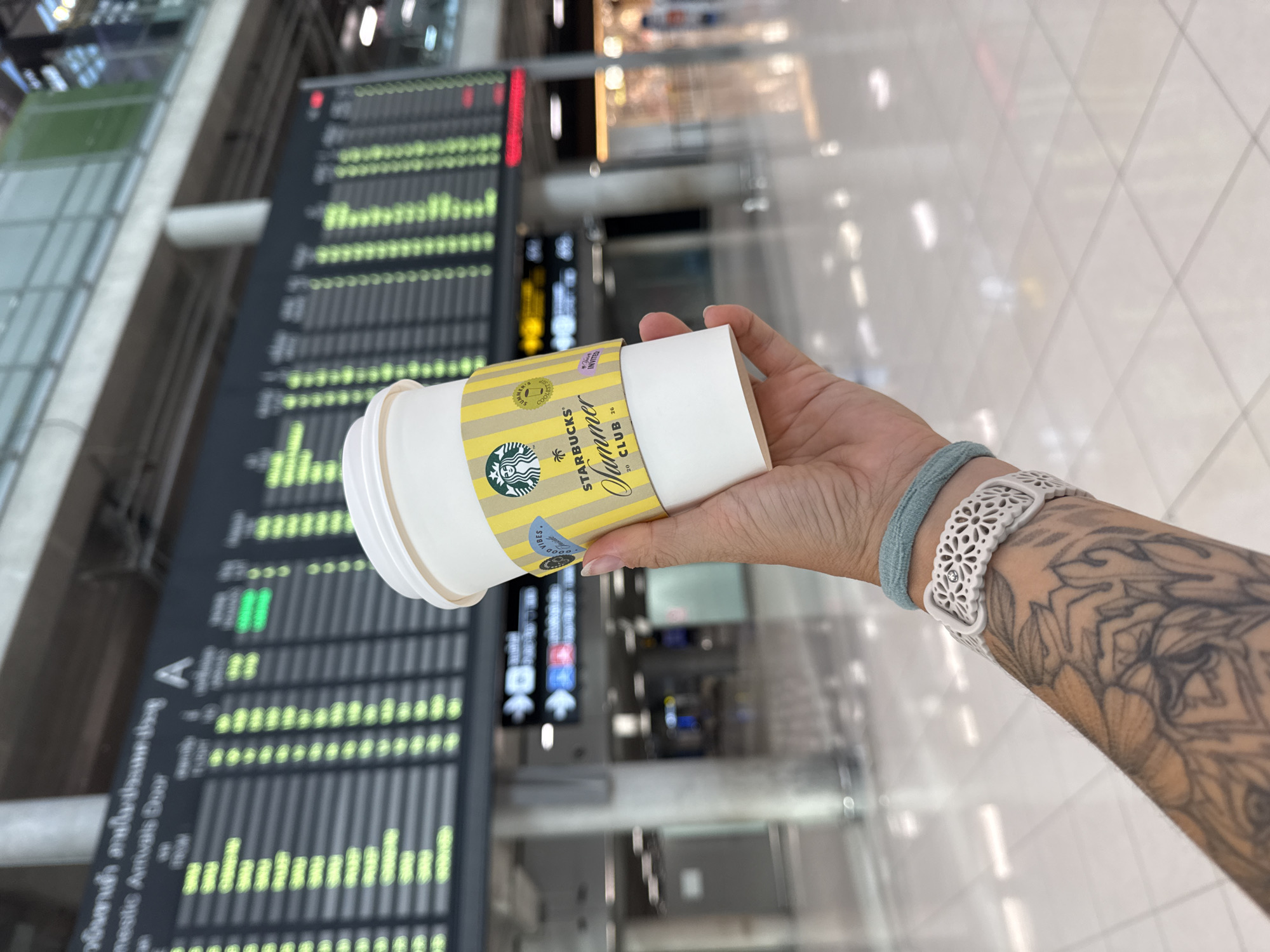 A person's hand is holding a Starbucks coffee cup in front of an airport departure board. The cup has a yellow and white sleeve with the Starbucks logo and "Summer Club" text. The person is wearing a light blue band and a white patterned bracelet on their wrist, and has tattoos on their arm. In the background, a large digital display board shows flight information, with many rows of text and green and red indicators. The floor appears to be tiled and reflective. The environment suggests an airport terminal in the city of Nong Prue Subdistrict, Thailand, given the Thai text visible on the departure board and the overall airport setting.