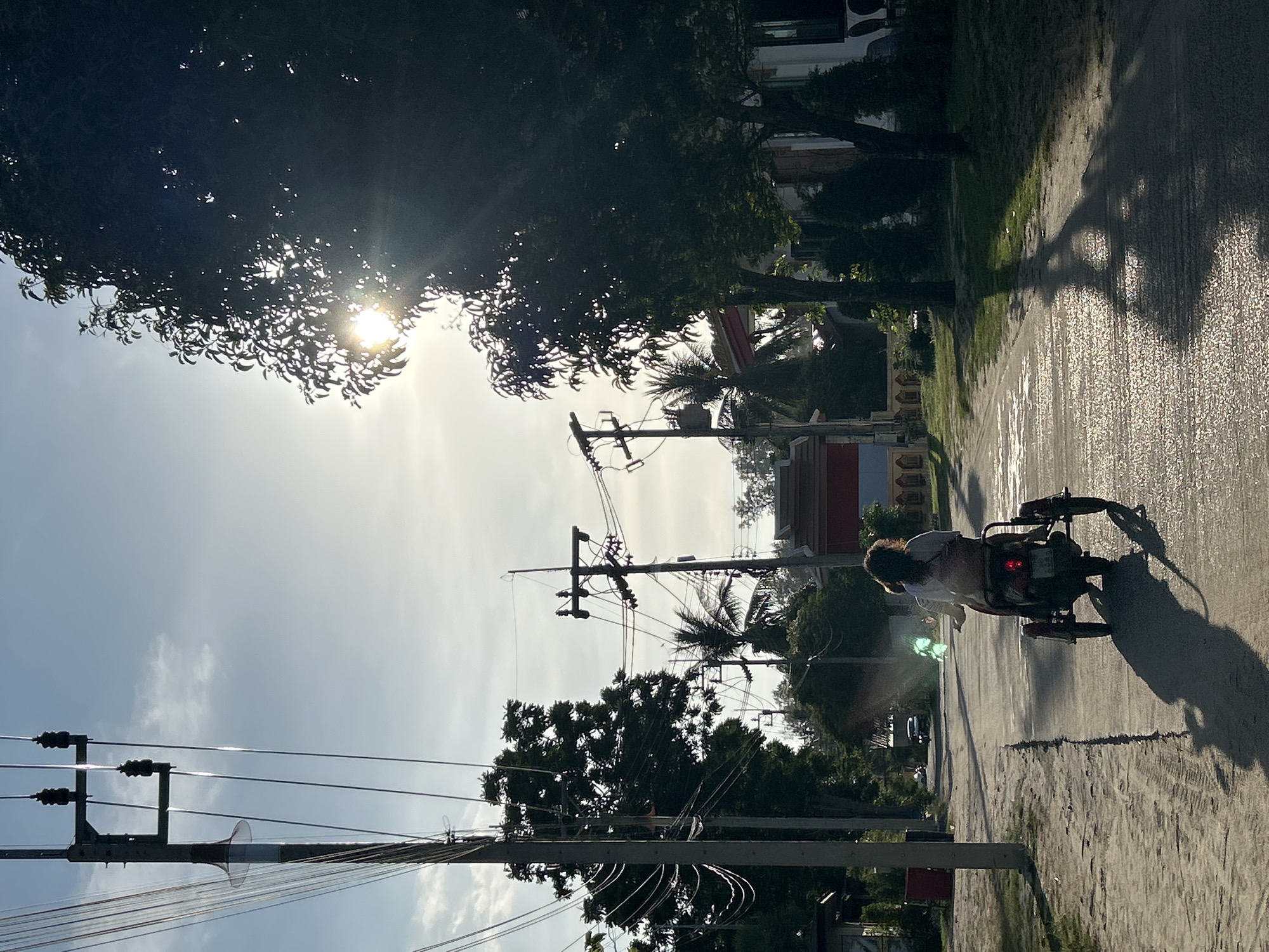 The image depicts a street scene in ตำบลเกาะพะงัน, Thailand, under a bright, sunlit sky obscured by trees. A utility pole and wires dominate the left side of the frame, stretching towards the upper-right. A person rides a motorcycle along the road, casting a long shadow. The road surface appears textured, possibly wet. Vegetation lines the sides of the road, with buildings visible in the background.
