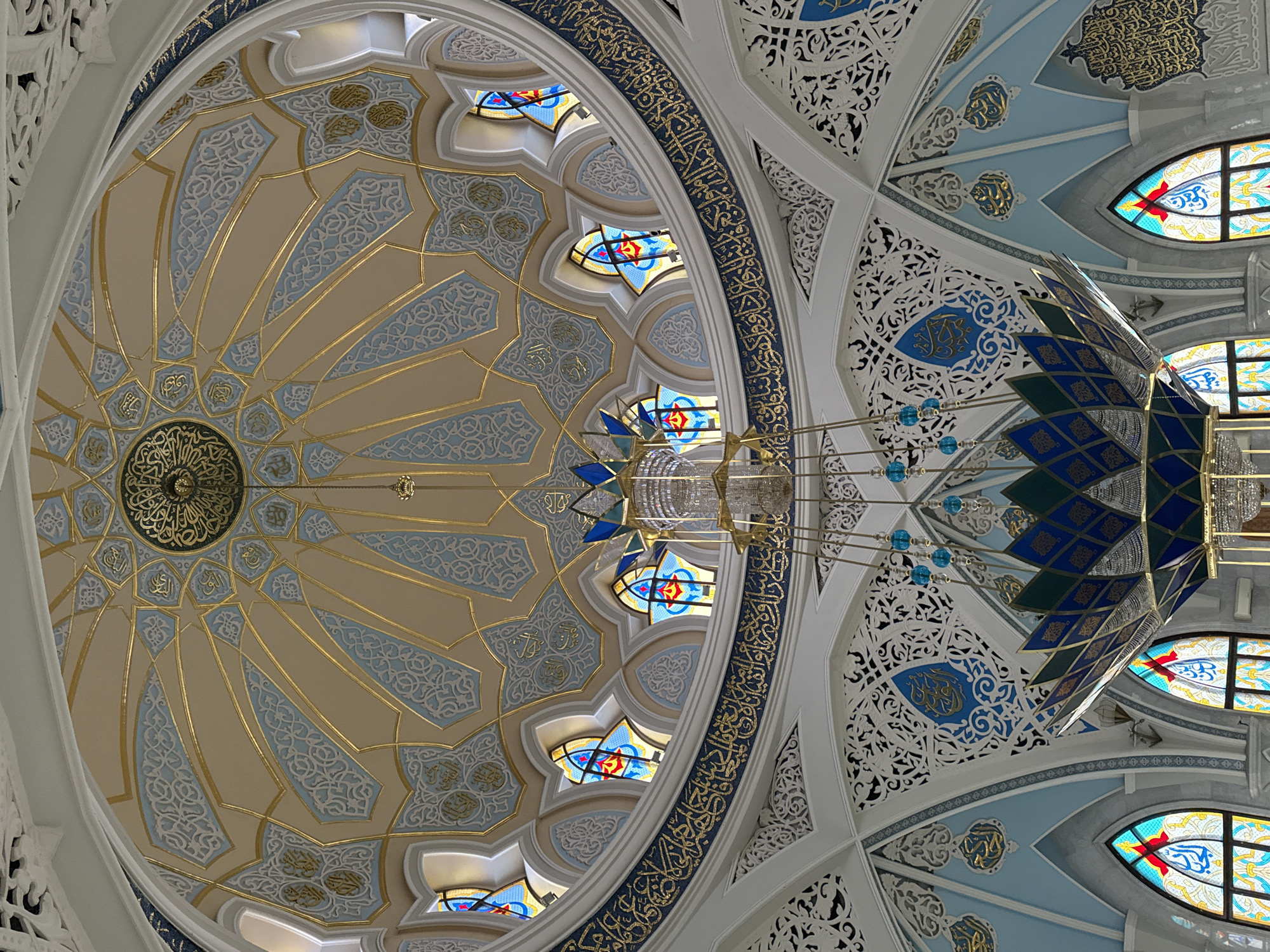 The image shows the interior of a mosque dome. The dome features a circular design with repeating geometric patterns in beige, light blue, and gold colors. Arabic calligraphy is also visible. Stained glass windows are incorporated into the design. A large chandelier with blue and green accents hangs from the center of the dome. The ceiling also features intricate white latticework and decorative blue accents. The image is likely taken in the city of Kazan, Russia, given the architectural style and religious symbolism.