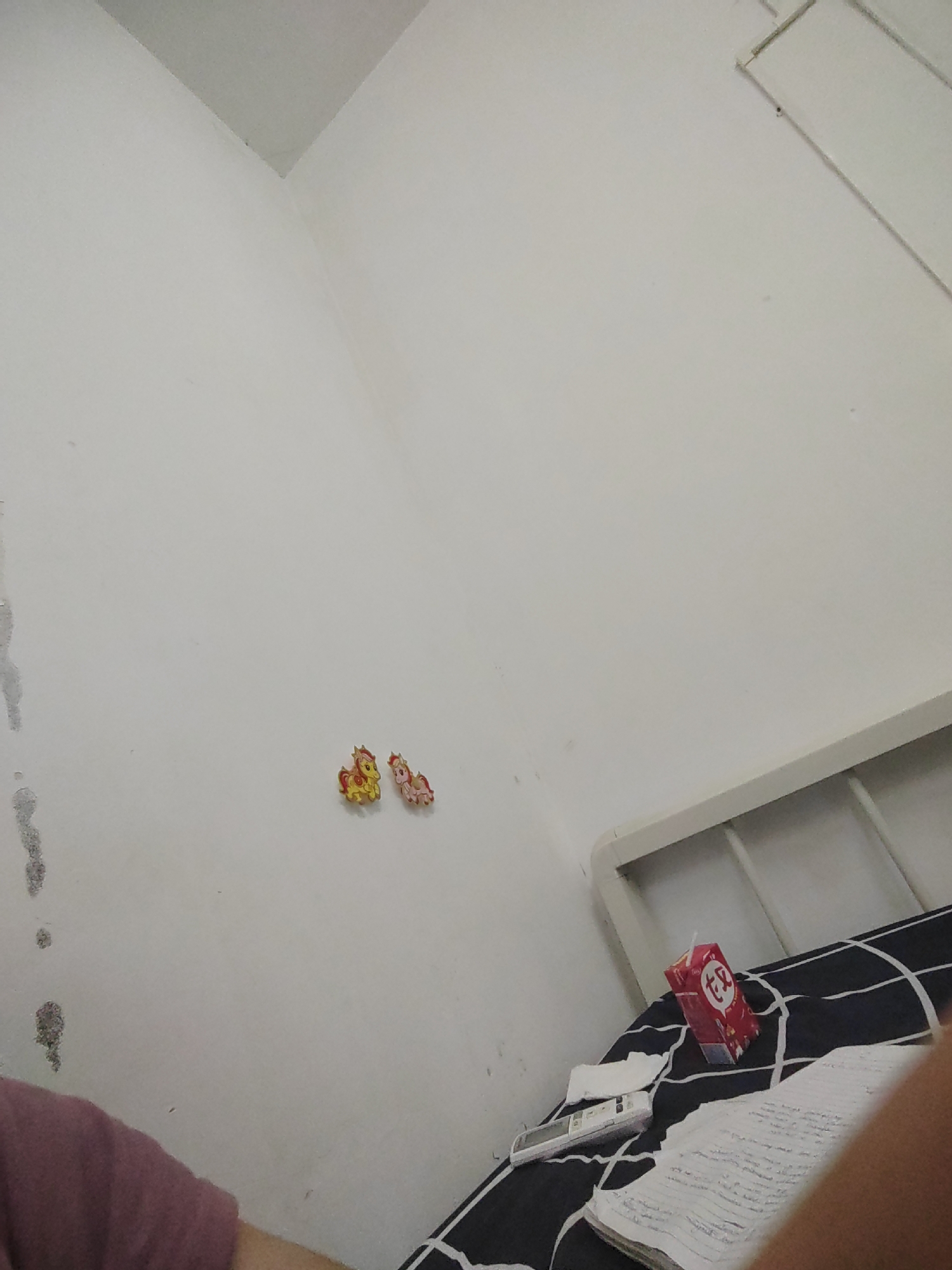 The image depicts a close-up view of a white wall in a room, with a portion of a bed visible in the lower right corner. Two small, colorful decorations resembling horses are affixed to the wall. On the bed, a red juice box with Chinese characters and a white straw is placed on a black and white striped comforter. A remote control and some papers are also on the bed. A person's arm, wearing a dark pink shirt, is visible in the lower left corner of the image. The environment appears to be a bedroom. The image is shot from a low angle, looking upwards.