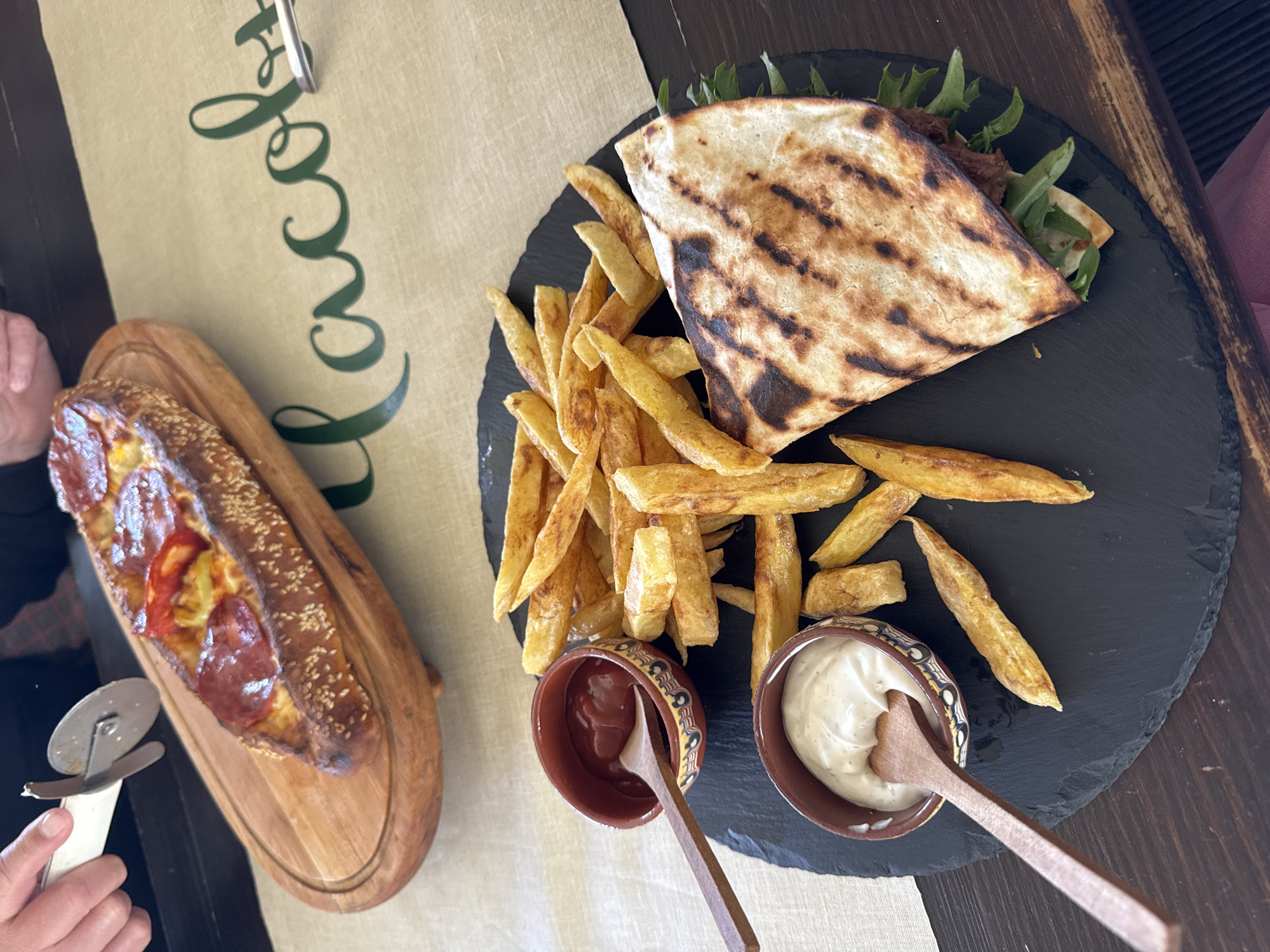 The image shows a top-down view of a table with food. On the right side, there's a black slate plate holding a folded pita sandwich, French fries, and two small bowls containing sauces (one red, one white), each with a small wooden spoon. To the left, a long, rectangular pizza is on a wooden board. Part of a person's hand is visible, holding a pizza cutter near the pizza. The table has a light-colored runner with dark green calligraphy-like lettering. The overall environment suggests a restaurant setting. The image does not provide explicit information on the location in the city of Blagoevgrad, Bulgaria.