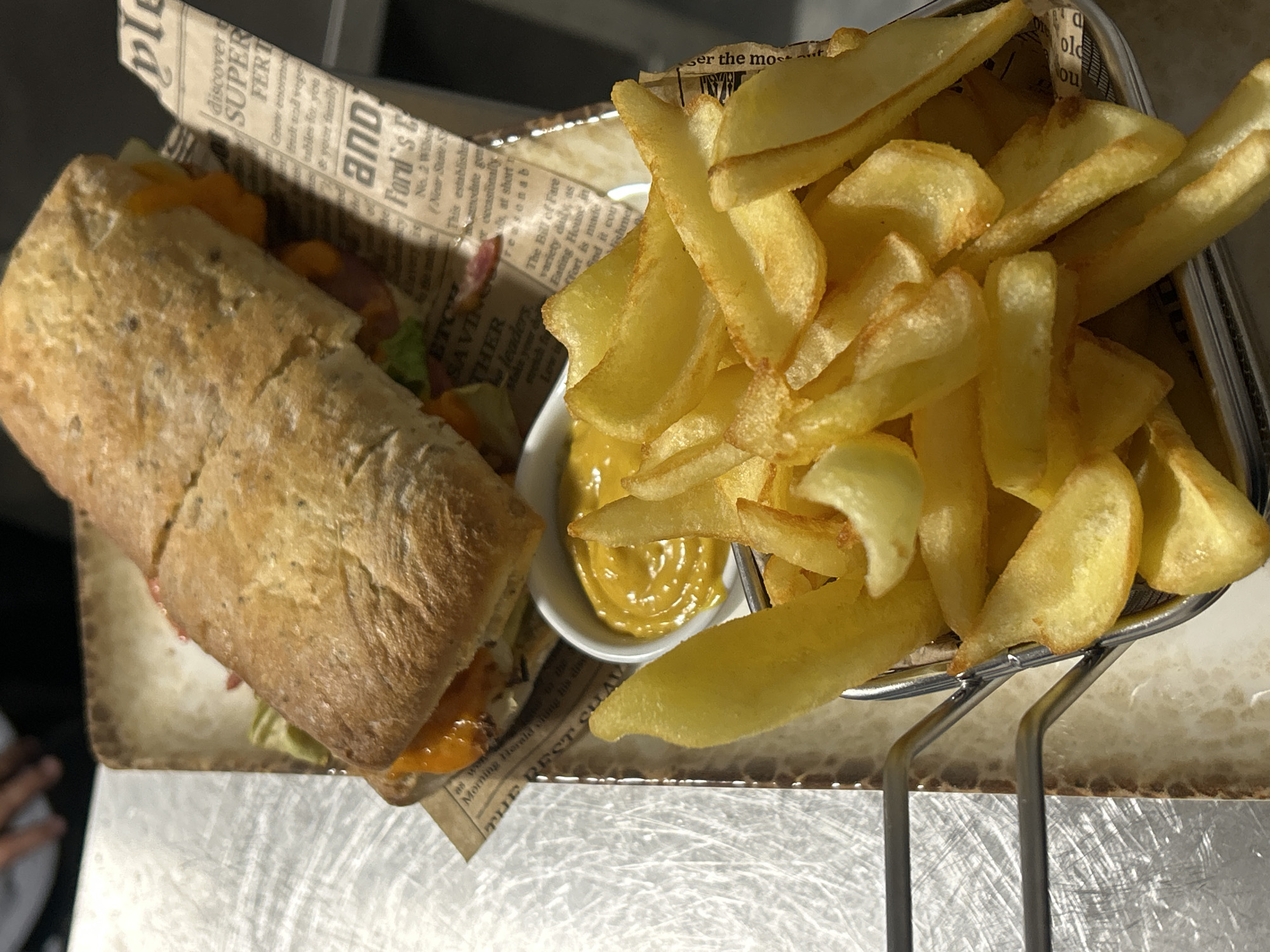 Here's a detailed description of the image:
The image shows a meal, likely a sandwich and fries, presented on a tray lined with newspaper-style print. The sandwich is a long baguette-style bread filled with visible layers of lettuce, a red ingredient (possibly meat or tomato), and what appears to be cheese. The fries are thick-cut and arranged in a metal serving basket. A small white bowl contains a yellow sauce, likely a dipping sauce for the fries. The meal is set on a stainless-steel surface, potentially a countertop in a restaurant or kitchen setting. The newspaper lining the tray has snippets of text, including mentions of "Ford's E," "Eating House," "the city of Burgas, Bulgaria "The Best".