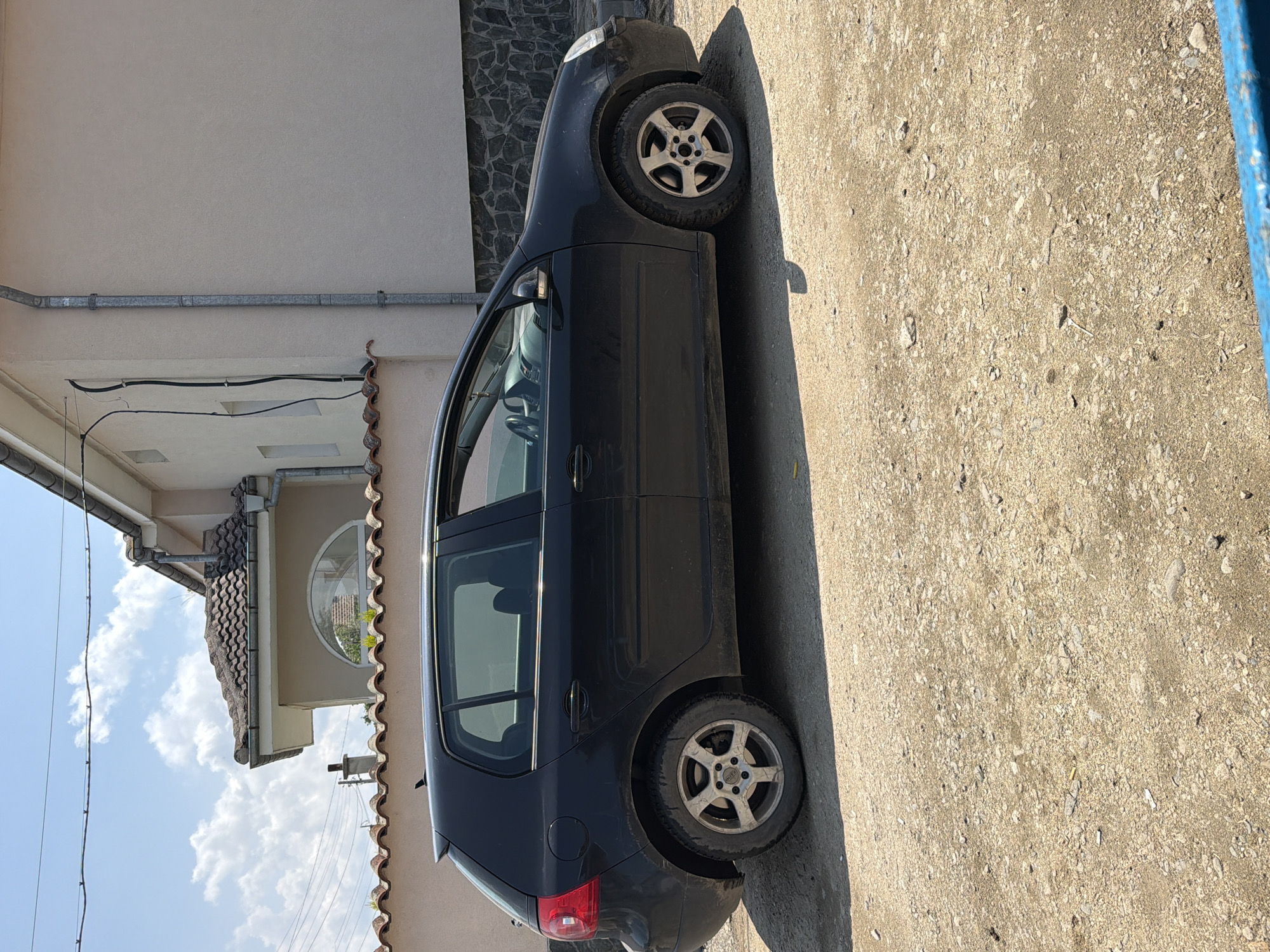 The image, taken in Dabene, Bulgaria, depicts a dark-colored (likely black or dark grey) five-door hatchback car parked on an unpaved, dirt, and gravel road. The car is positioned with its passenger side visible, facing away from the viewer. Its silver alloy wheels have an Audi logo on the hubcaps, and the car appears somewhat dusty. A long shadow cast by the vehicle suggests bright mid-day or afternoon sunlight.
To the left of the car is a building with a beige plastered facade. A section of its corner features a decorative stone veneer. The building has a brown tiled roof, beneath which a distinct circular window is visible, reflecting greenery and the sky. A silver-colored pipe runs horizontally along the upper part of the wall, with black electrical cables attached below it. Overhead, the sky is clear blue with scattered white cumulus clouds. No people are visible, and no specific activity is taking place other than the car being parked. The scene conveys a quiet, outdoor setting, possibly in a residential or rural area.