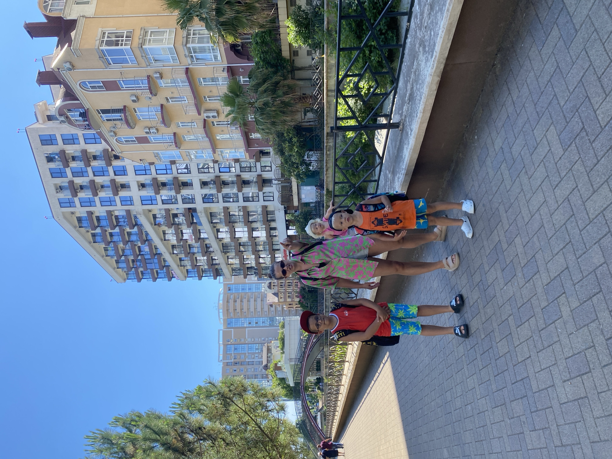 The media file shows a group of children on a paved walkway next to a railing, with a large, modern building in the background. The building is light yellow and beige with many balconies and windows. Palm trees and other greenery are visible around the building and in the distance. The sky is clear and blue, indicating a sunny day.
There are at least three children visible. One child in the foreground wears a red t-shirt, glasses, and blue and green patterned shorts. Next to them is another child wearing an orange t-shirt and light-colored shorts. Further back, a child in a pink and green patterned outfit is visible. The children appear to be posing for the camera and seem happy and relaxed.
The scene appears to be in a resort or urban area, possibly near the coast, given the palm trees and the style of the building. The paved walkway suggests a public area like a promenade or a park. The bright sunlight and clear sky suggest it is daytime, likely mid-morning or afternoon. The overall mood is cheerful and summery. There is a red hat on one child and some text on their shirt. The text on the orange shirt is mostly obscured but includes the word "SKATE". The red t-shirt has text that says "RE:AY" and other illegible text.