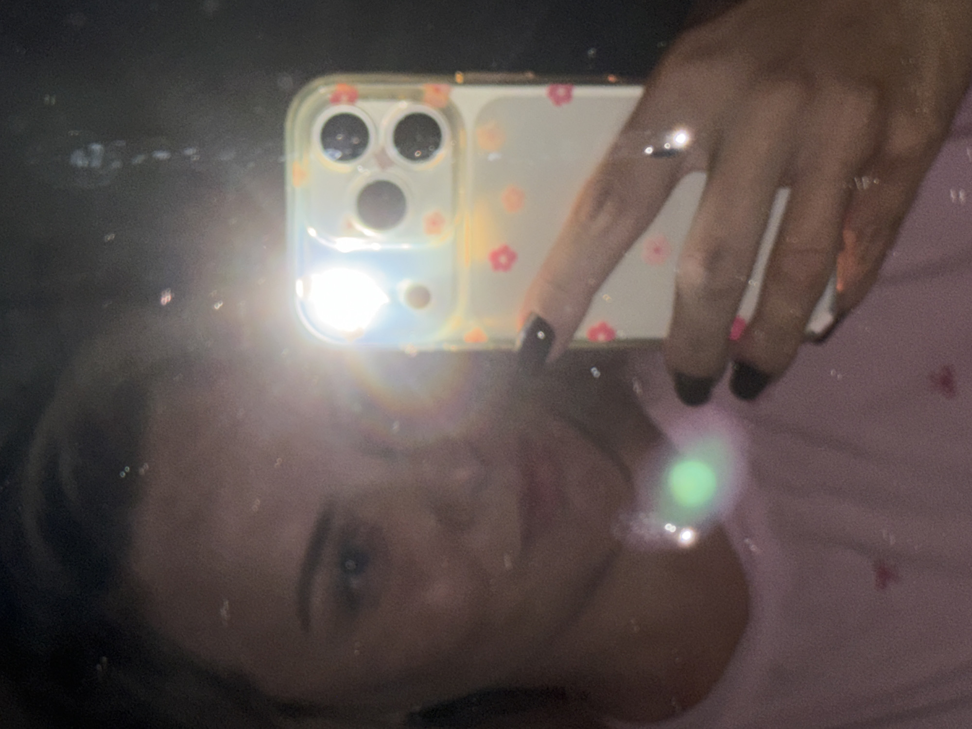 A slightly blurry, overhead selfie shows a young woman in a pink shirt holding an iPhone with a clear case decorated with small pink and orange flowers. The phone's camera flash is on, casting a bright light across the image. The woman is looking at the phone's reflection in what appears to be a mirror, with her face visible from the forehead to the chin. Her hair is light brown and pulled back. Her nails are painted black. The background is dark and appears to be a room. There are some water spots or dust particles on the reflective surface, creating small bright flares in the image. The woman is in the city of Votkinsk, Russia.