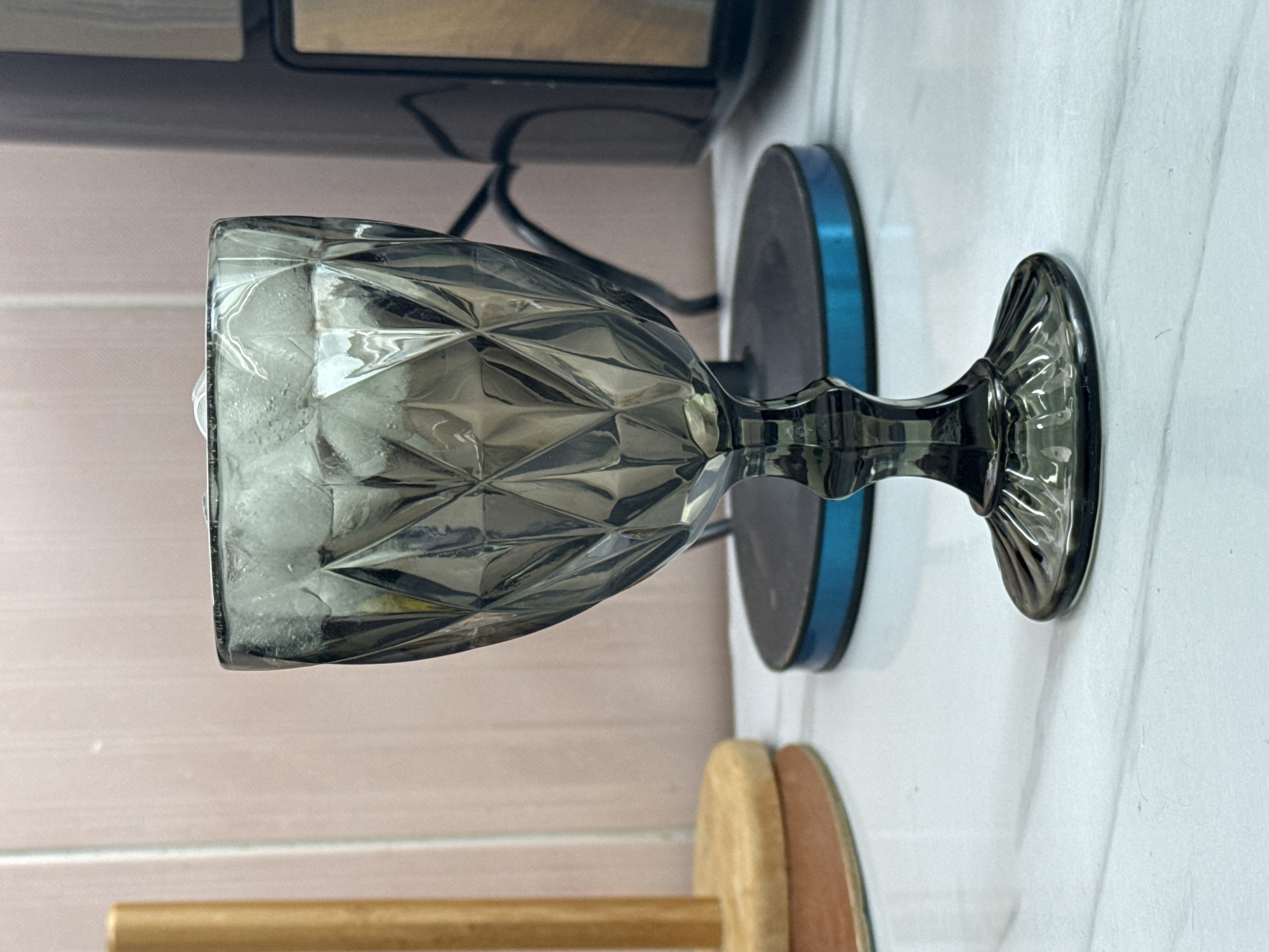 The scene, located in Khimki, Russia, presents a still life of a dark grey, faceted glass goblet containing ice cubes. The goblet is centrally positioned on a white countertop exhibiting subtle grey veining. To the left, a portion of a wooden paper towel holder, consisting of a circular base and a vertical dowel, is visible. To the right of the goblet, a dark-colored kitchen appliance, possibly a coffee maker, is partially visible, featuring a prominent blue ring around its base. The background consists of a light pink, textured wall. No individuals, direct actions, or interactions are depicted.
