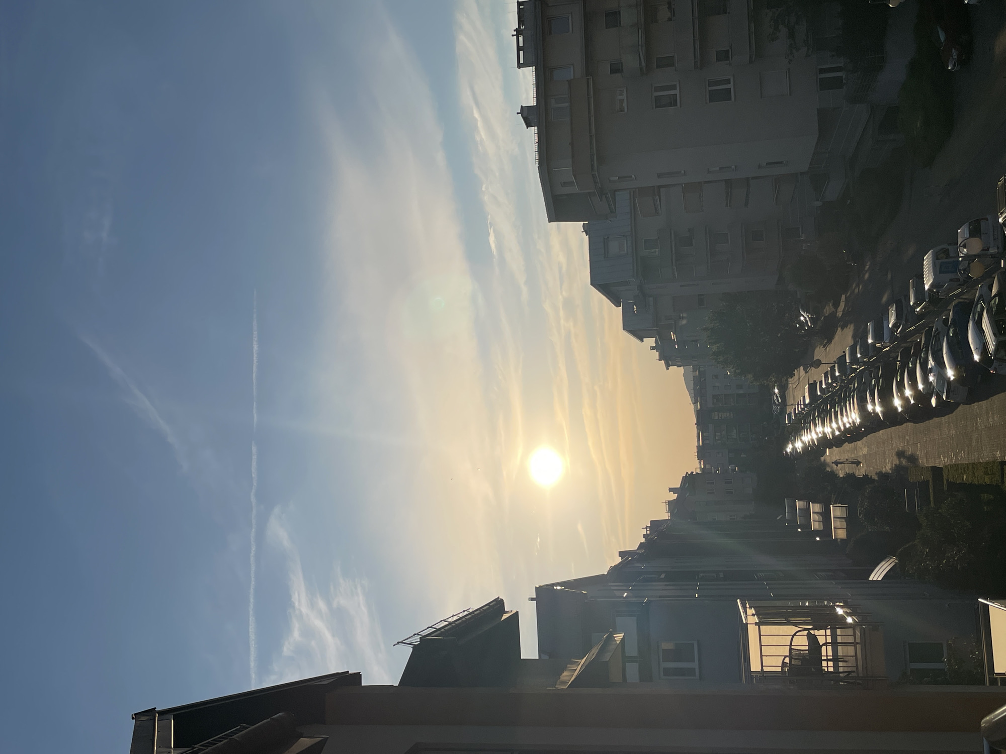 The image depicts a city street scene at sunset, viewed from an elevated perspective. The sun, a bright orb, is setting low in the sky, casting a warm, golden glow and creating lens flare. The sky is a mix of clear blue and streaks of wispy clouds, with contrails from aircraft visible.
Dominating the frame are several multi-story buildings, likely apartments or offices, characterized by their angular architecture and numerous windows. On the right side of the image, a long row of parked cars lines the street, their surfaces reflecting the sunlight. Trees and greenery are interspersed between the buildings and along the street, adding a touch of nature to the urban environment.
The overall impression is one of a peaceful evening in a built-up area, with the setting sun imbuing the scene with a sense of calm and warmth. There are no people or discernible activities taking place. The time of day is clearly sunset due to the position and color of the sun and the lighting conditions. No specific location cues are visible, and no text is present in the image. The image appears to be taken with a smartphone camera, given the slight distortion and the presence of lens flare.