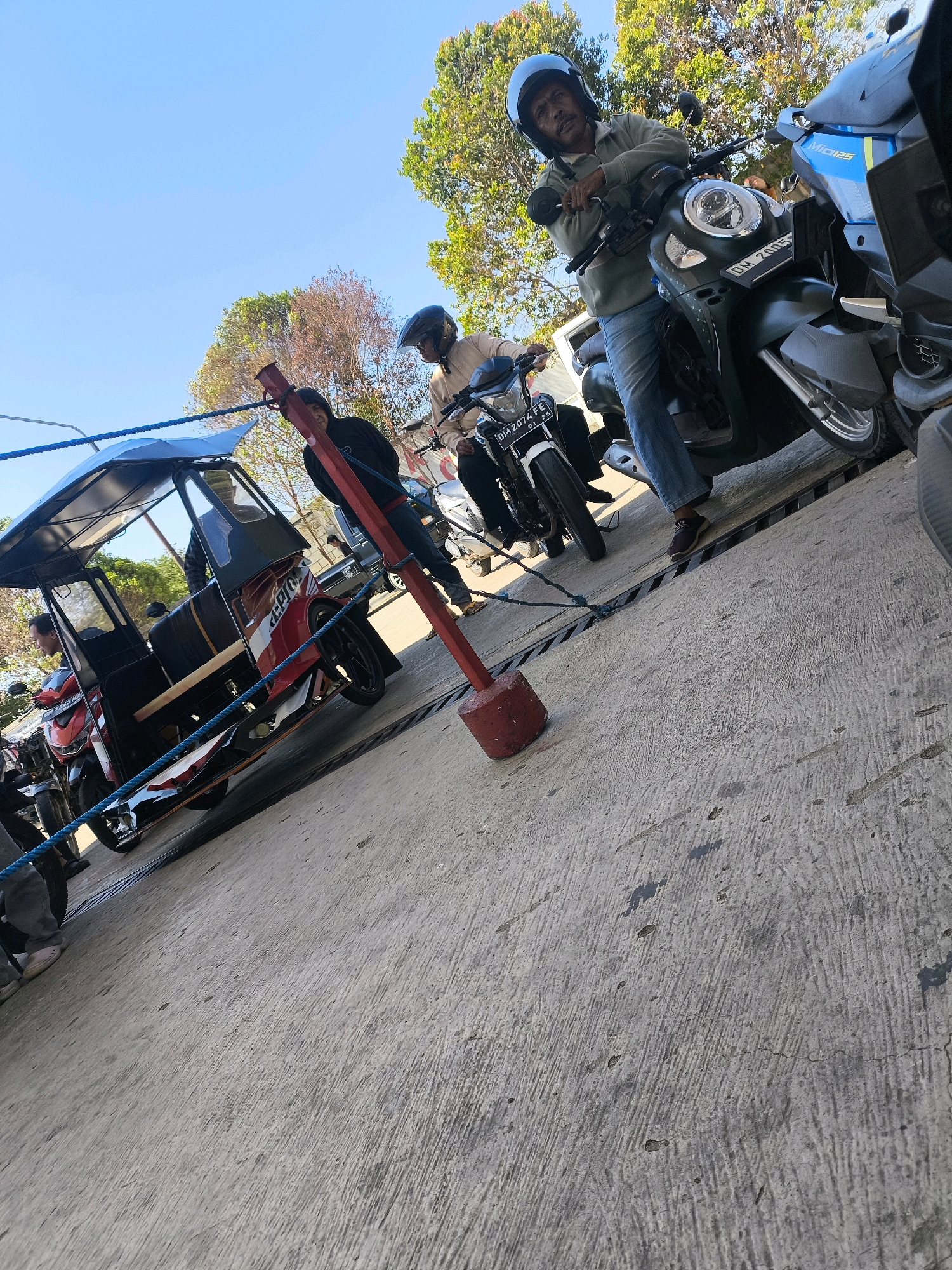 In Kwandang, Indonesia, a group of people and vehicles are gathered on a concrete surface under a clear blue sky with trees in the background. A man wearing a helmet and a light-colored jacket is seated on a dark green motorcycle. To his right, another motorcycle with a blue and yellow design is visible. In the foreground, a red and white three-wheeled vehicle, possibly a bajaj or motor-rickshaw, is parked. Two other individuals are near the three-wheeled vehicle, with one appearing to be seated inside. Another person in a black shirt is standing near the three-wheeled vehicle. A blue rope is stretched across the scene, attached to a red and white post. The ground is a rough, grey concrete with visible cracks and some dark patches. The overall scene suggests a transportation hub or a waiting area for these vehicles.