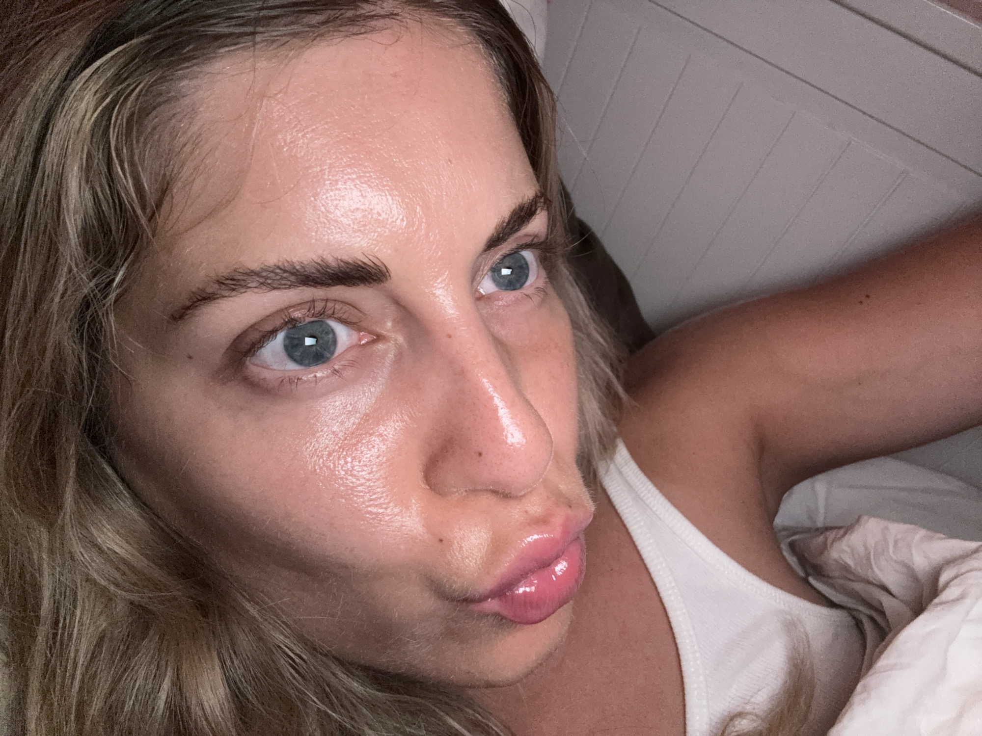 A close-up, low-angle, indoor shot shows a woman with blonde hair, blue eyes, and pink lips pouting. She is wearing a white tank top and is partially covered by a blanket. The background appears to be a wall with horizontal panels. The image does not provide any information about the location in Paris, France.