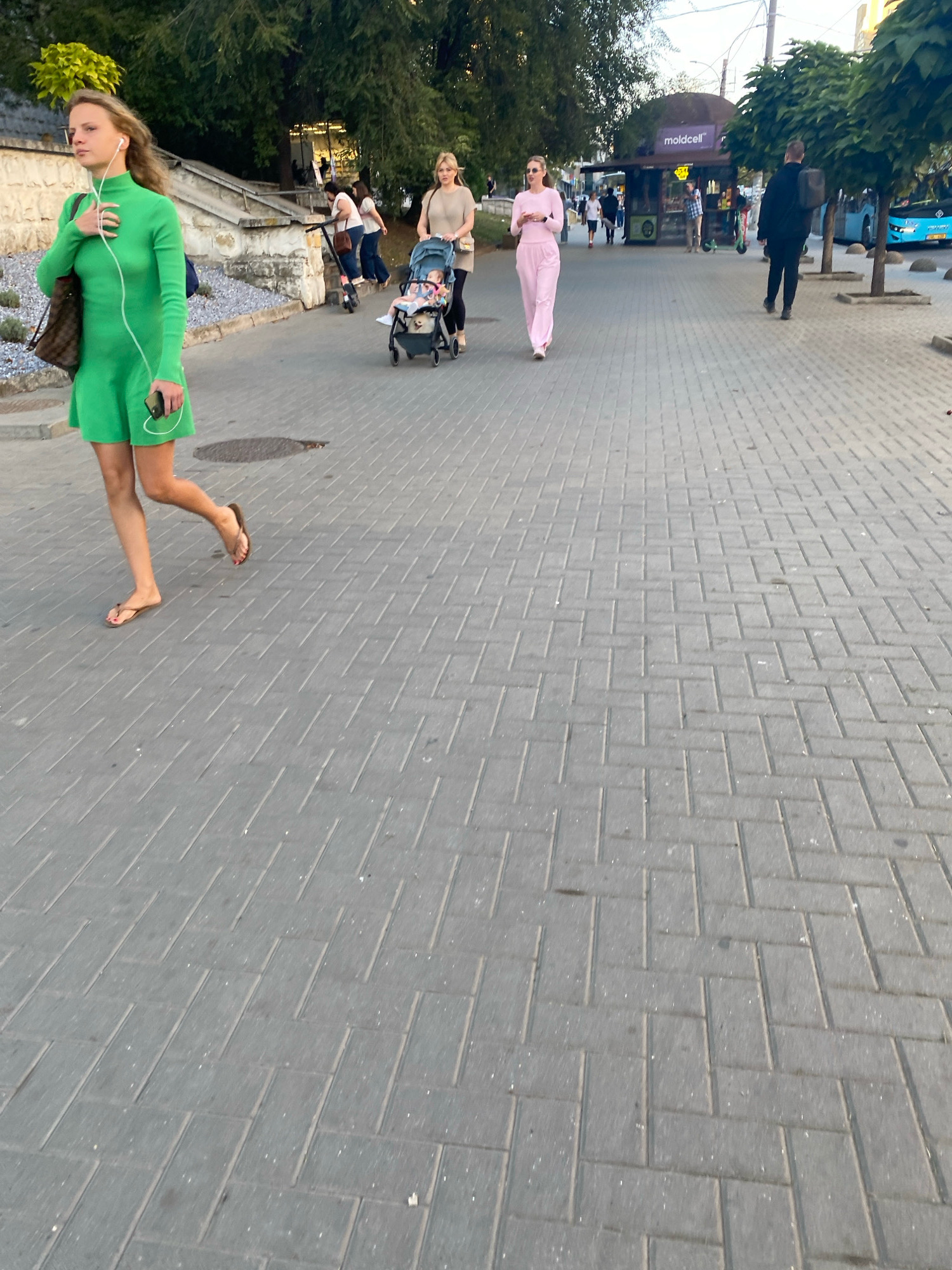 A woman in a green dress walks on a pedestrian sidewalk made of interlocking paving stones. She is wearing headphones and holding a smartphone. Behind her, a woman pushes a stroller with a baby. Further back, other people are visible on the sidewalk, as well as a kiosk labeled "moldcell". Trees line the sidewalk, and a blue bus is seen in the background. The scene appears to be outdoors during the day, with a soft, natural light. This photograph was taken on Ștefan cel Mare și Sfânt Boulevard in Chișinău, Moldova.