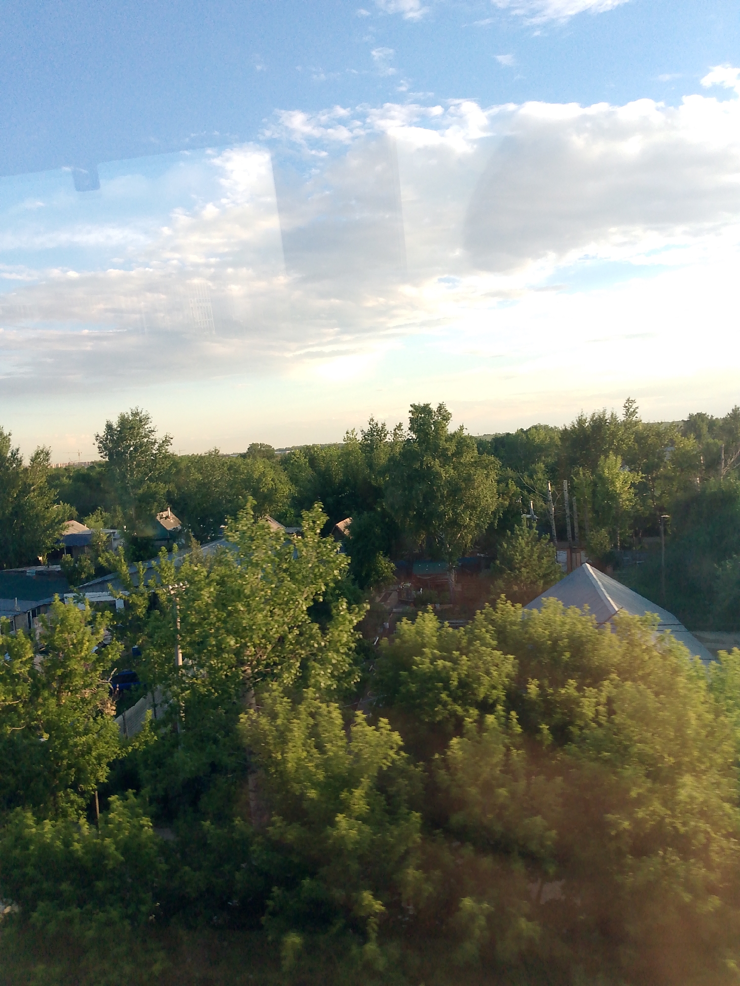 This image depicts a landscape scene, likely taken from an elevated vantage point, possibly a window. The foreground is dominated by lush green trees with sunlight dappling through their leaves, suggesting a warm, possibly late afternoon or early evening, time of day.
In the mid-ground, several small houses or cottages with visible roofs are nestled amongst more trees. Some of these structures appear to have chimneys. Beyond the houses, a dense forest stretches towards the horizon, with some taller structures, possibly cranes or distant buildings, visible in the far distance, hinting at development or a city on the outskirts.
The sky is a clear blue with scattered, fluffy white clouds, indicating pleasant weather. The overall atmosphere is serene and peaceful, conveying a sense of rural or suburban tranquility. There are no people or explicit activities visible, only the static elements of the landscape. No text is discernible in the image. The scene is consistent with a residential area in or around Astana, Kazakhstan, with its characteristic blend of greenery and development.