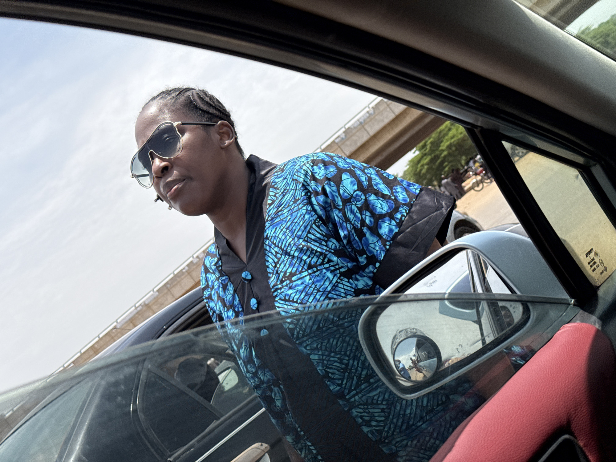 An eye-level shot from inside a car captures a woman leaning out of the passenger window during bright daylight. The woman, positioned centrally, wears dark aviator-style sunglasses and a vibrant blue and black patterned top or jacket, featuring distinct button detailing on the neckline. Her hair is styled in braids. She looks forward and slightly downwards, her expression obscured by the sunglasses.
The car's interior elements frame the scene, including the dark roof lining, the window frame, and a textured red panel, possibly an armrest, in the bottom right corner. The exterior side mirror is prominent, reflecting a blurred street scene with distant figures, including what appears to be a person on a bicycle or motorbike, and buildings under a sunny sky. The side mirror itself has a sticker or label that clearly reads "TOYOTA" and "MARK II JZX100," indicating the car's make and model. Another faint sticker within the car's window seems to include the word "MAXIMUM."
In the background, under a bright, slightly overcast sky, a large concrete bridge structure stretches across the upper half of the image, suggesting an overpass or elevated road. Green foliage from trees is visible below and to the right of the bridge. The scene depicts a dynamic moment in what appears to be an urban or semi-urban environment in Galadimawa Village, Nigeria, with the woman possibly observing or interacting with something outside the vehicle.