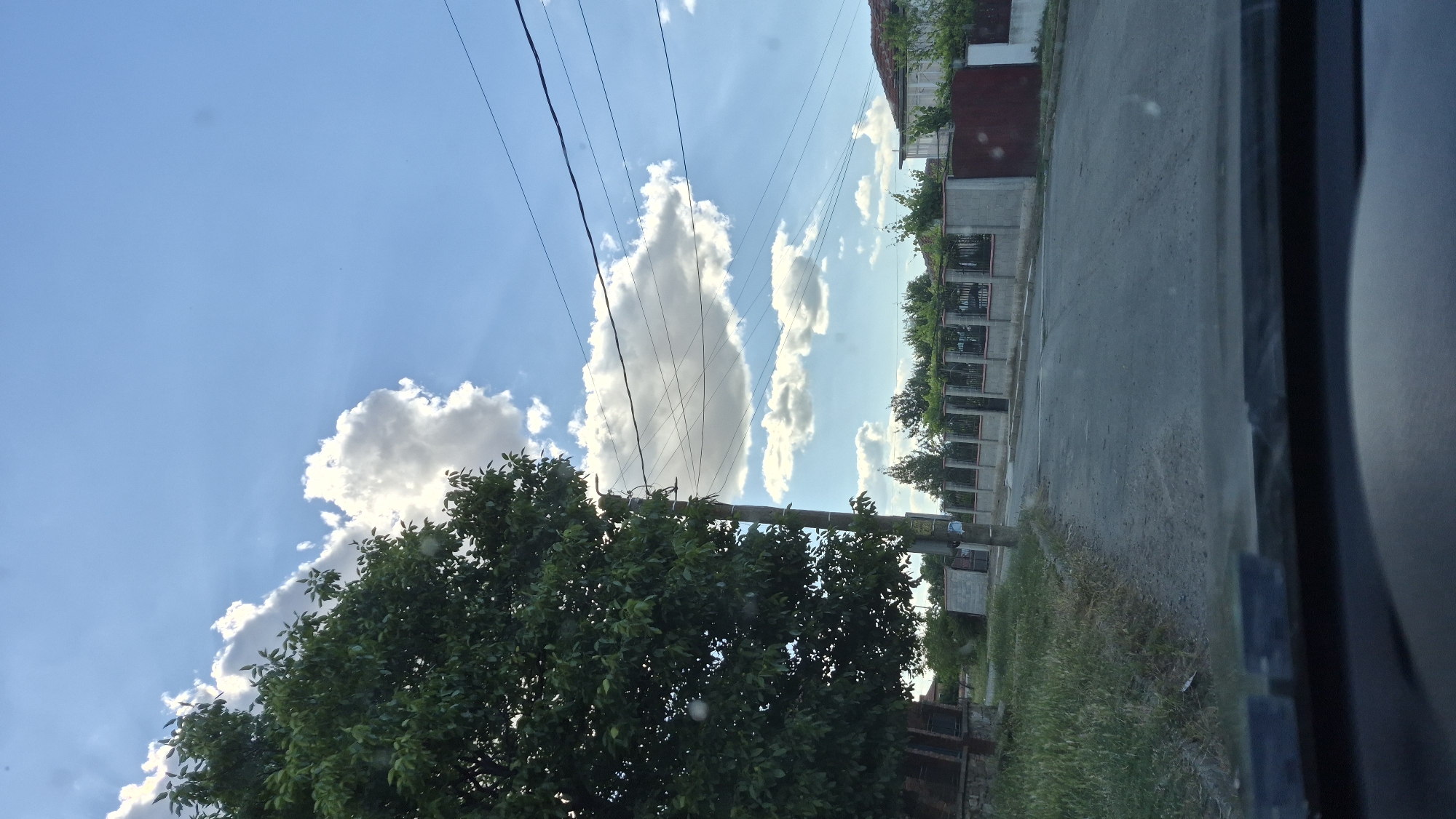 The image depicts a street scene in a rural or suburban area. In the foreground, the dashboard of a vehicle is visible, suggesting the photo was taken from inside a car. The road ahead is unpaved and appears to be a dirt or gravel surface, winding slightly to the right.
To the left, a large, leafy green tree dominates the scene, its branches extending towards the sky. Adjacent to the tree, there's a glimpse of a brick structure. A utility pole with numerous wires strung between it and other unseen poles stands prominently in the middle ground, near the tree.
The background features a row of houses and fences, indicating a residential area. The fences are constructed with concrete pillars and metal railings, some sections are solid concrete. Beyond the fences, more trees and vegetation are visible. The sky is a clear blue, punctuated by several fluffy white clouds, with rays of sunlight visible breaking through them. The overall impression is a sunny day in a quiet, possibly developing, neighborhood.
