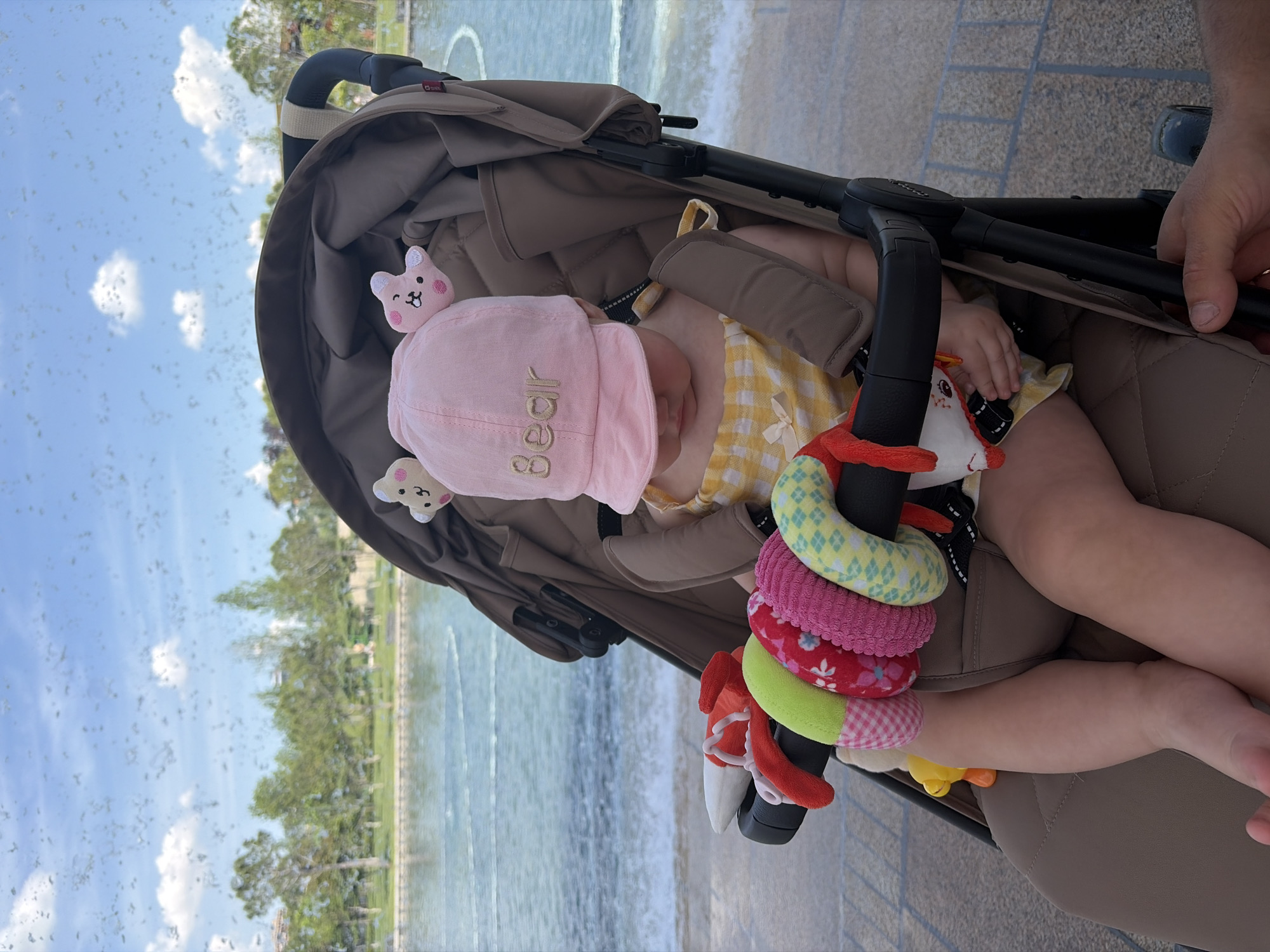 The image displays an infant situated in a beige stroller, observed through a surface bearing multiple water droplets. The infant wears a pink bucket hat with "Bear" embroidered on the front, complemented by a yellow and white checkered top. The child is engaged with a white plush toy, part of a multicolored spiral activity toy affixed to the stroller's bumper bar. An adult's right hand grasps the stroller frame on the lower right. The background reveals a wide body of water with visible ripples, bordered by a distant treeline under a clear blue sky with sparse clouds. This setting is identified as Krasnodar Park (Galitsky Park) in Krasnodar, Russia.