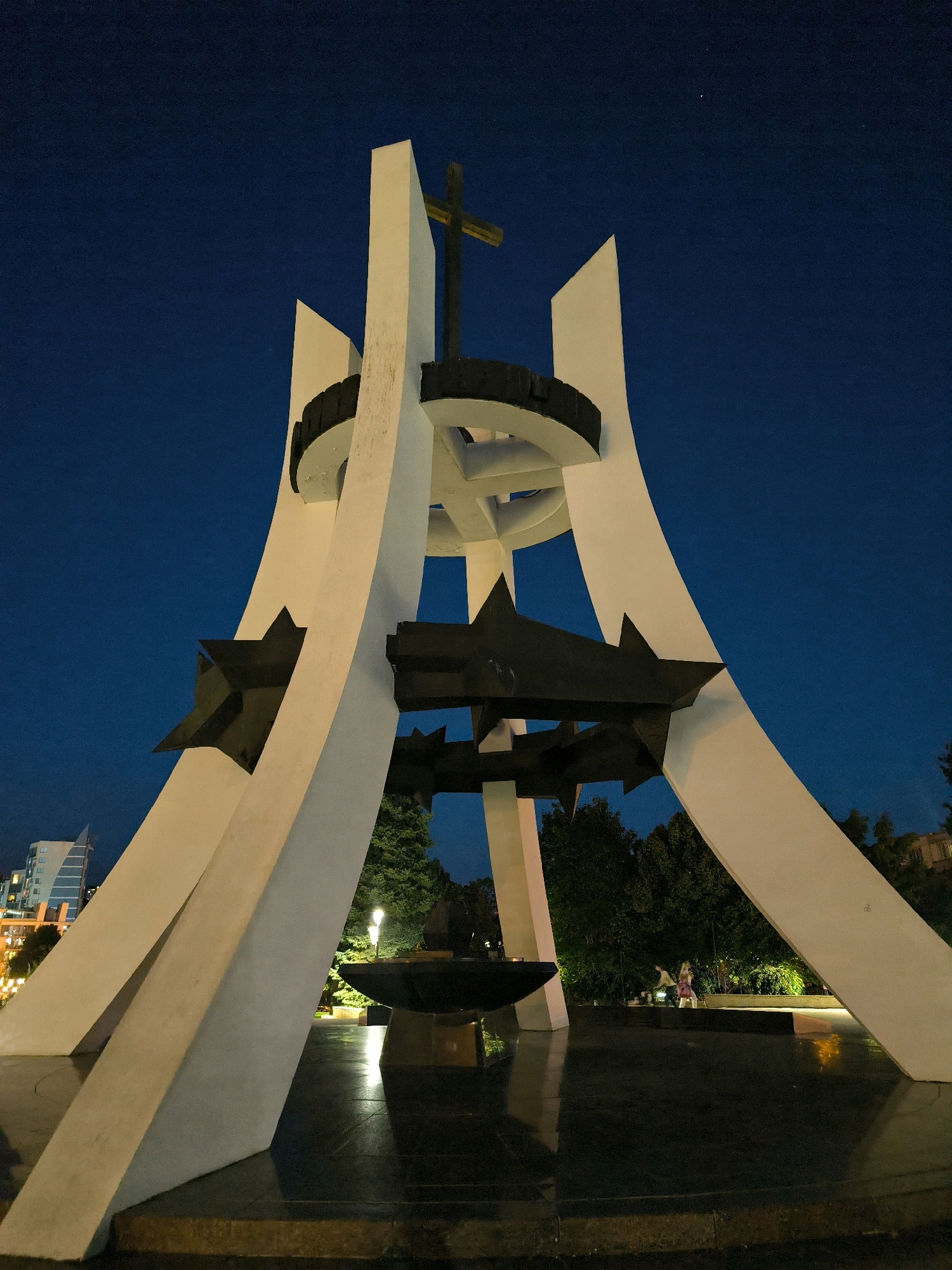 The image depicts a monumental structure at dusk in Chișinău, Moldova. The monument, made of white concrete and dark metal, features abstract architectural elements with a cross at its apex. Three dark, star-shaped objects are affixed to the structure's metallic rings, which are situated between white pillars. Below the monument, a dark, reflective surface, likely polished stone or water, mirrors the surrounding lights. In the background, trees and distant buildings, illuminated by artificial lights, are visible against a deep blue twilight sky. Two individuals can be seen in the mid-ground, standing near the monument. A single streetlight illuminates the area.
