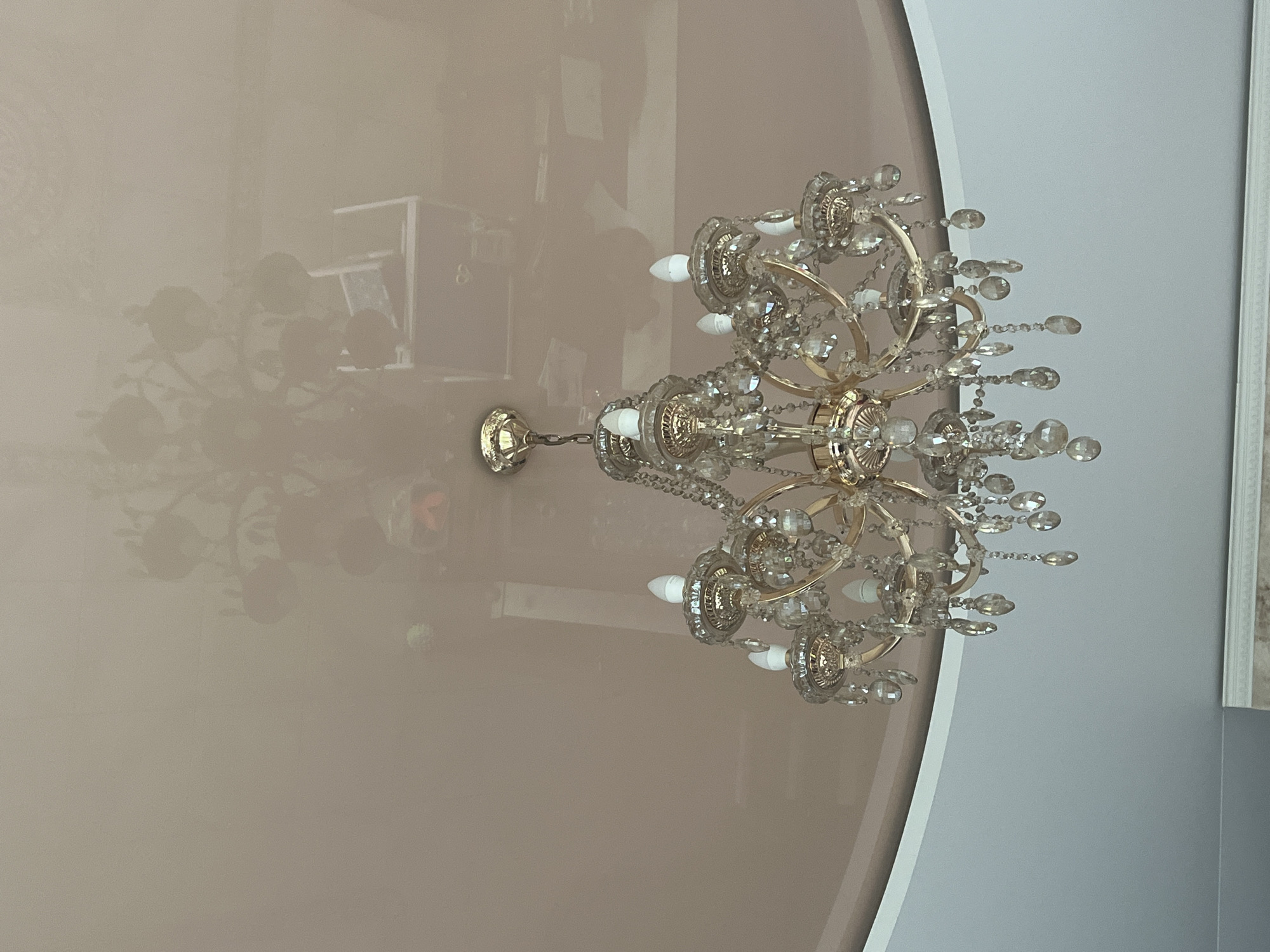 A crystal chandelier hangs from a reflective, light pink ceiling. The chandelier is made of gold-toned metal and features multiple arms adorned with clear crystal prisms and teardrop-shaped pendants. Each arm holds a white, LED light bulb. The ceiling reflects the chandelier and other objects, including what appears to be furniture and possibly a computer monitor, in a distorted manner. The image is taken from a low angle, looking upwards. The location is specified as Vladimirovka, Russia.