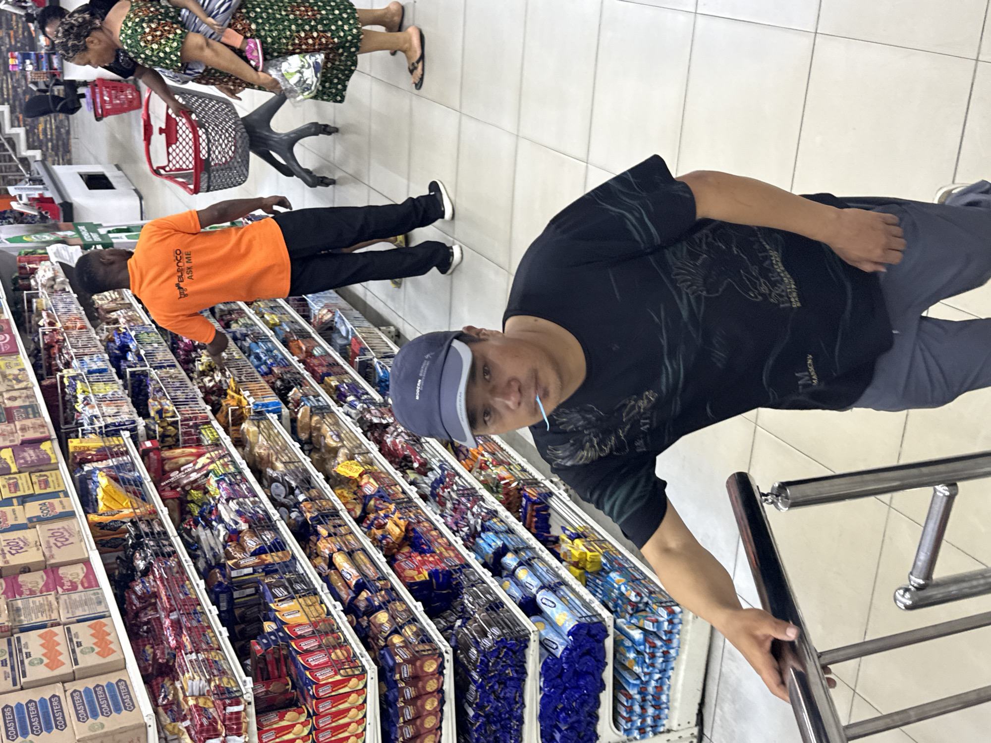 Here is a detailed description of the image:
**Location:** Appears to be inside a grocery store in Ado, Nigeria.
**Environment:** Aisle within a grocery store, with rows of shelves stocked with various packaged snacks and goods. The floor is tiled, and there's a metallic handrail visible, seemingly used for navigation by shoppers.
**People:**
* A man in a dark patterned t-shirt, grey trousers, and a blue cap is holding onto the handrail. He has a toothpick or something similar in his mouth.
* A person wearing an orange t-shirt with "BLENCO ASK ME" printed on the back.
* Partial views of other shoppers including one with a shopping basket and another wearing a green patterned dress, carrying a child.
**Objects:**
* Shelves are filled with packaged goods, including snacks, biscuits, and drink mixes.
* Packaging details visible: "COASTERS", "OLD", "SODA", "MANSONY", and "MODERN"
* Shopping basket of red color
**Actions/Interactions:**
* People are navigating the aisle to shop.
* The man in the orange t-shirt appears to be browsing or stocking the shelves.
**In summary:** The image depicts an average shopping scene inside a grocery store in Ado, Nigeria.