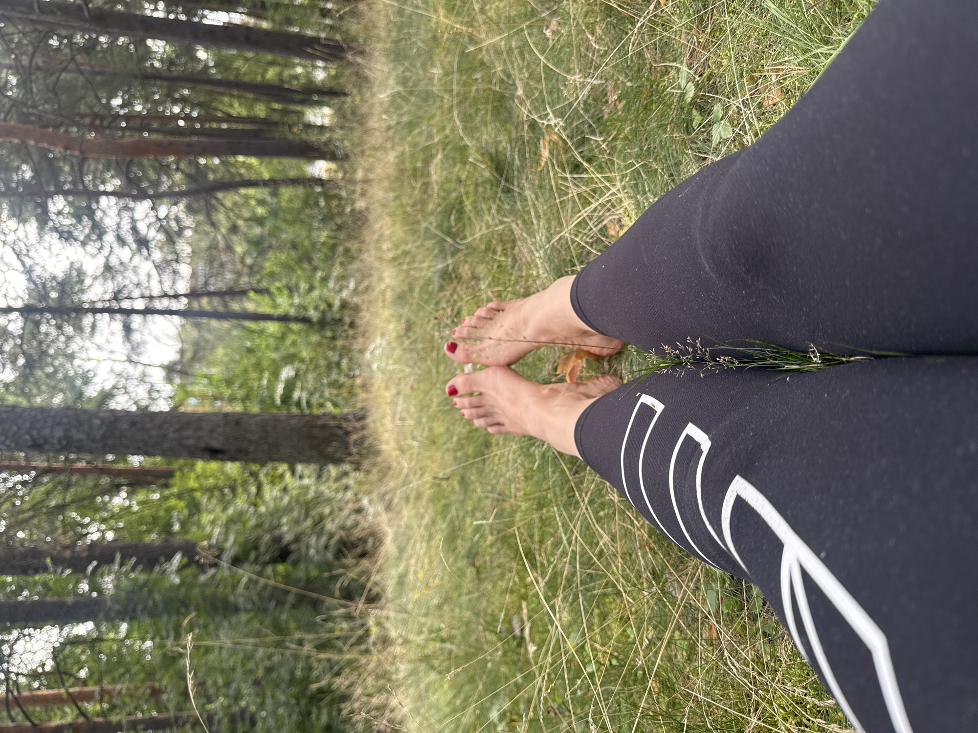 The image shows a first-person perspective of a person lying on their back in a grassy, wooded area. The person is wearing black leggings with a white geometric pattern. Their bare feet, with red nail polish, are visible in the foreground, resting on the grass. The background consists of trees and foliage, suggesting a natural outdoor setting, possibly a park or forest. There is no indication of the city of Sosnoviy Bor, Russia, within the image itself.