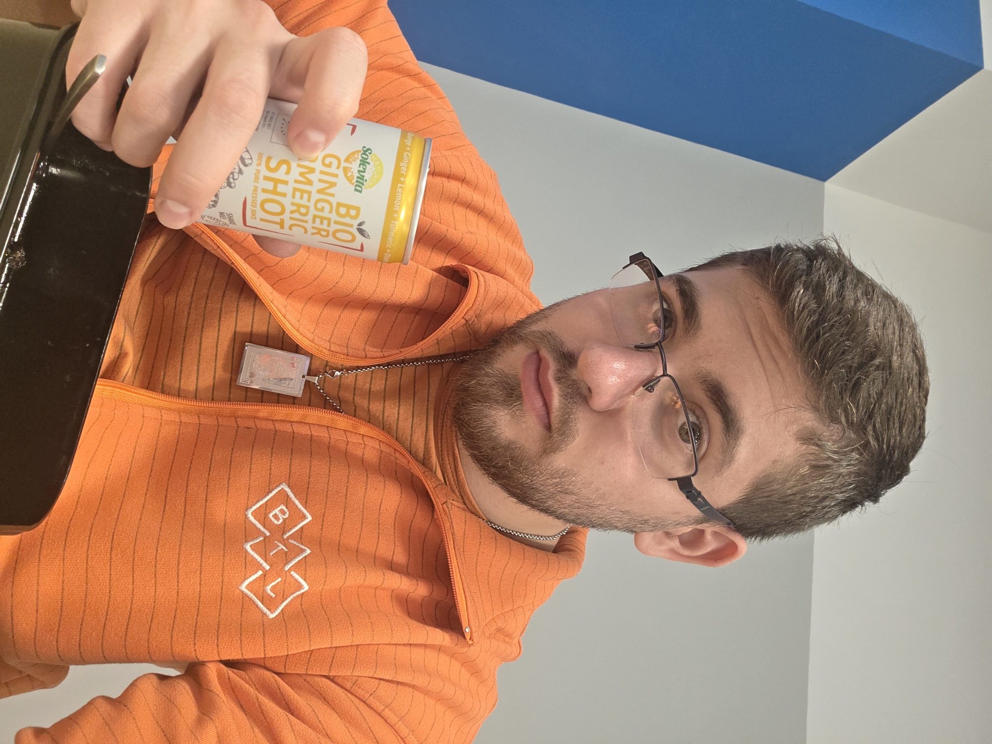 A fair-skinned man with short brown hair, a beard, and glasses wears an orange zip-up jacket with a white logo. He holds a black object in one hand and a "Solevita Bio Ginger Tumeric Shot" can in the other. The background is a white and blue wall. It is impossible to determine if the photo was taken in Plovdiv, Bulgaria.