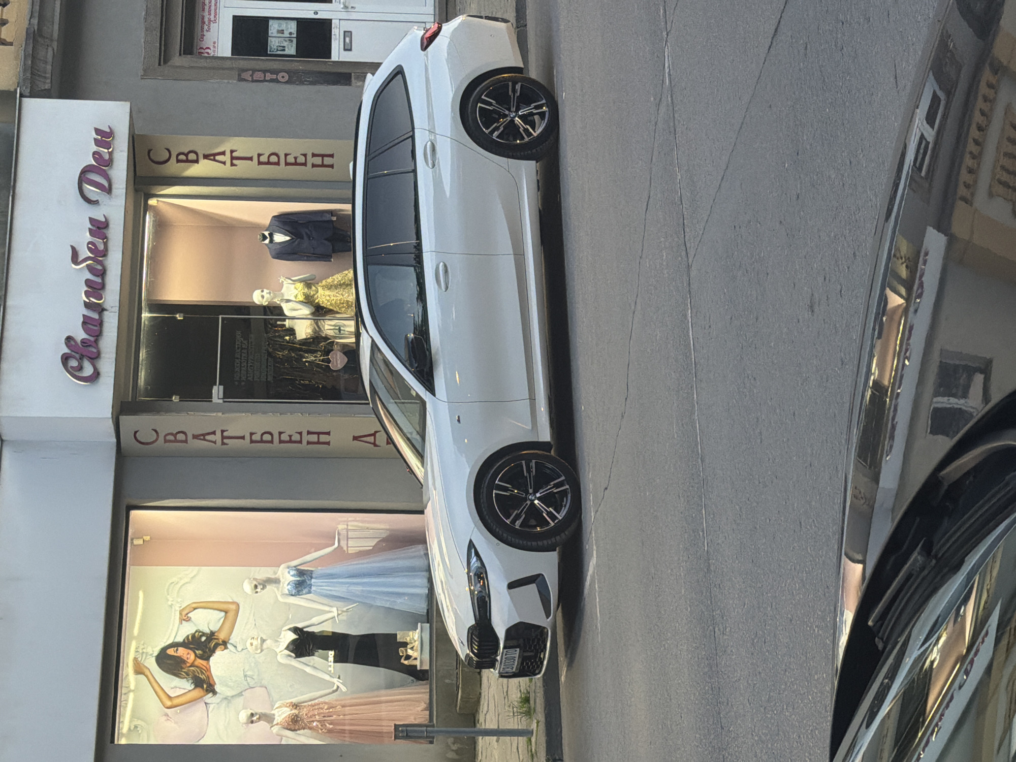The image displays a street scene in Shumen, Bulgaria, featuring a white modern car parked next to a storefront. The car, possibly a BMW, is oriented vertically in the frame and exhibits multi-spoke black and silver wheels. Its front license plate, "S S8039TO," is clearly visible. The car's polished surface reflects buildings and the street from the opposite side, indicating a bright, sunny daytime setting.
The storefront belongs to a clothing shop, likely specializing in formal wear for weddings and proms. A large sign above the entrance prominently displays "Сватбен Ден" (Svatben Den), translating to "Wedding Day." This text, "CBATBEH," is also seen vertically on the shop's glass window. Inside the display windows, several mannequins showcase various formal garments, including a dark suit, a light blue gown, a patterned gold dress, a dark formal dress, and a rose-gold colored dress. A large pictorial graphic of a smiling woman also features within the display. Another Cyrillic word, "АБИТУРИЕНТС," translating to "Prom Graduates," is visible on the window, suggesting the store caters to prom events.
Above the main shop sign, faint Cyrillic text can be deciphered, including "ABTO" (Auto) and other less clear phrases such as "Скулпторые меде" and "Кобода-вленска," which may belong to another business or a previous tenant of the building. The street surface is paved with asphalt, and the overall impression is that of a bustling commercial area.