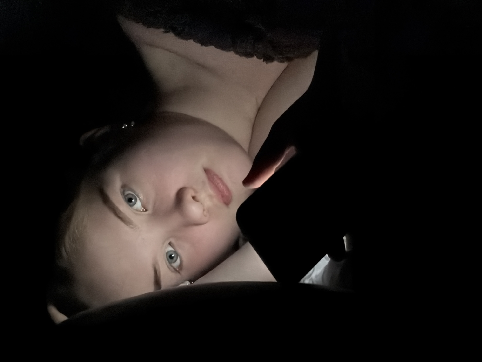 A female individual is depicted in a close-up, horizontal composition within a dark indoor environment, identified as being in the city of Chaikovsky, Russia. The primary illumination originates from a smartphone held in her right hand, casting an upward glow on her face. She possesses fair skin, light-colored hair, and blue eyes, with a noticeable blush on her cheeks. A small, light-colored stud earring is visible on her left earlobe. Her attire includes a black garment with lace detailing, evident on the strap over her right shoulder. Her left arm is bent, with the hand resting out of frame or obscured by darkness near her head. Her gaze is directed towards the camera. The background is rendered in complete darkness, obscuring all other environmental details.