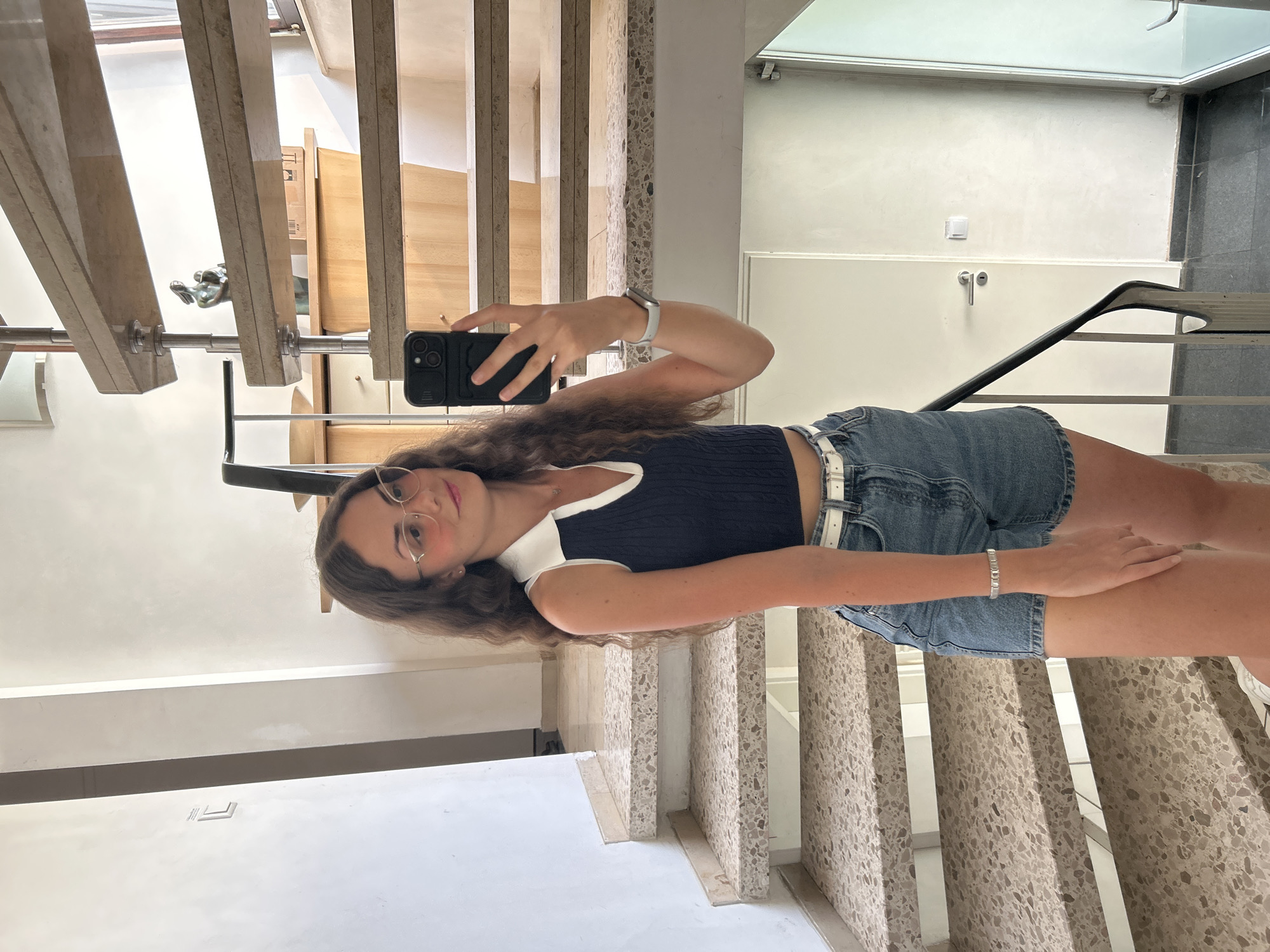 A young woman with long, wavy brown hair and glasses is standing on a staircase, taking a mirror selfie with her phone. She is wearing a dark blue sleeveless top with a white collar and collar opening, and denim shorts with a white belt. She has a bracelet on her left wrist. The staircase is made of beige stone with dark gray risers and a black metal railing. The background shows a modern interior with white walls, a door, and wooden furniture. The lighting is bright and natural.