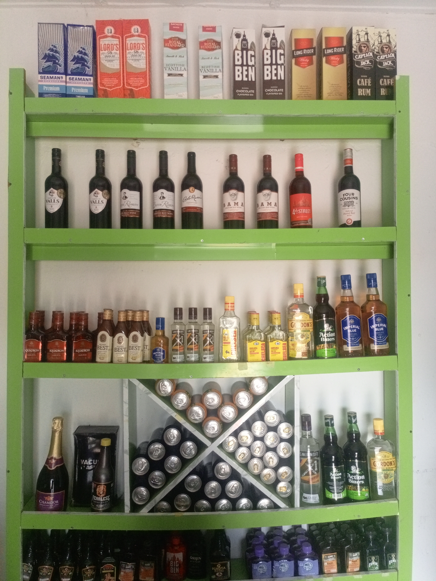 A well-lit interior shot captures a multi-tiered green metal shelf unit filled with various beverages and canned goods. The scene is likely a retail establishment or storage area, possibly a shop in the city of Jos, Nigeria, given the arrangement and product types.
The top shelf displays boxed items, including "Seamans Premium Schnapps" in blue packaging, "Lord's Gin London Dry" in red and white boxes, and smaller boxes labeled "Scottish Vanilla" and "Royal Standard." Further to the right, there are boxes for "Big Ben" flavored gin and whiskey, "Long Rider" whiskey, and "Captain Jack Cafe Rum."
The second shelf from the top features several dark glass bottles, mostly wine, with labels such as "Valls," "Baron Romero," and "4th Street." There is also a bottle labeled "Four Cousins Sweet Red Wine."
The third shelf from the top holds an assortment of smaller bottles and some full-sized bottles. These include "Squadron" rum, "Best" bottles, and "Imperial Blue" whiskey. A bottle of "Gordon's Dry Gin" and "Action Bitters" are also visible.
The fourth shelf contains a bottle of "Chamdor" sparkling grape juice, a black box with yellow lettering reading "VACUUM FAST," and a bottle with a lion's head logo labeled "FEARLESS." To the right, a section of the shelf is filled with stacked aluminum cans, arranged in a diamond pattern, appearing to be beverages.
The bottom shelf contains rows of dark bottles, some with colorful labels. Several small, dark bottles with purple caps are prominent, along with other smaller liquor bottles.
The background is a plain white wall, and the lighting suggests an indoor environment, possibly with artificial light. No people or animals are present in the image. The focus is entirely on the organized display of products on the shelf.