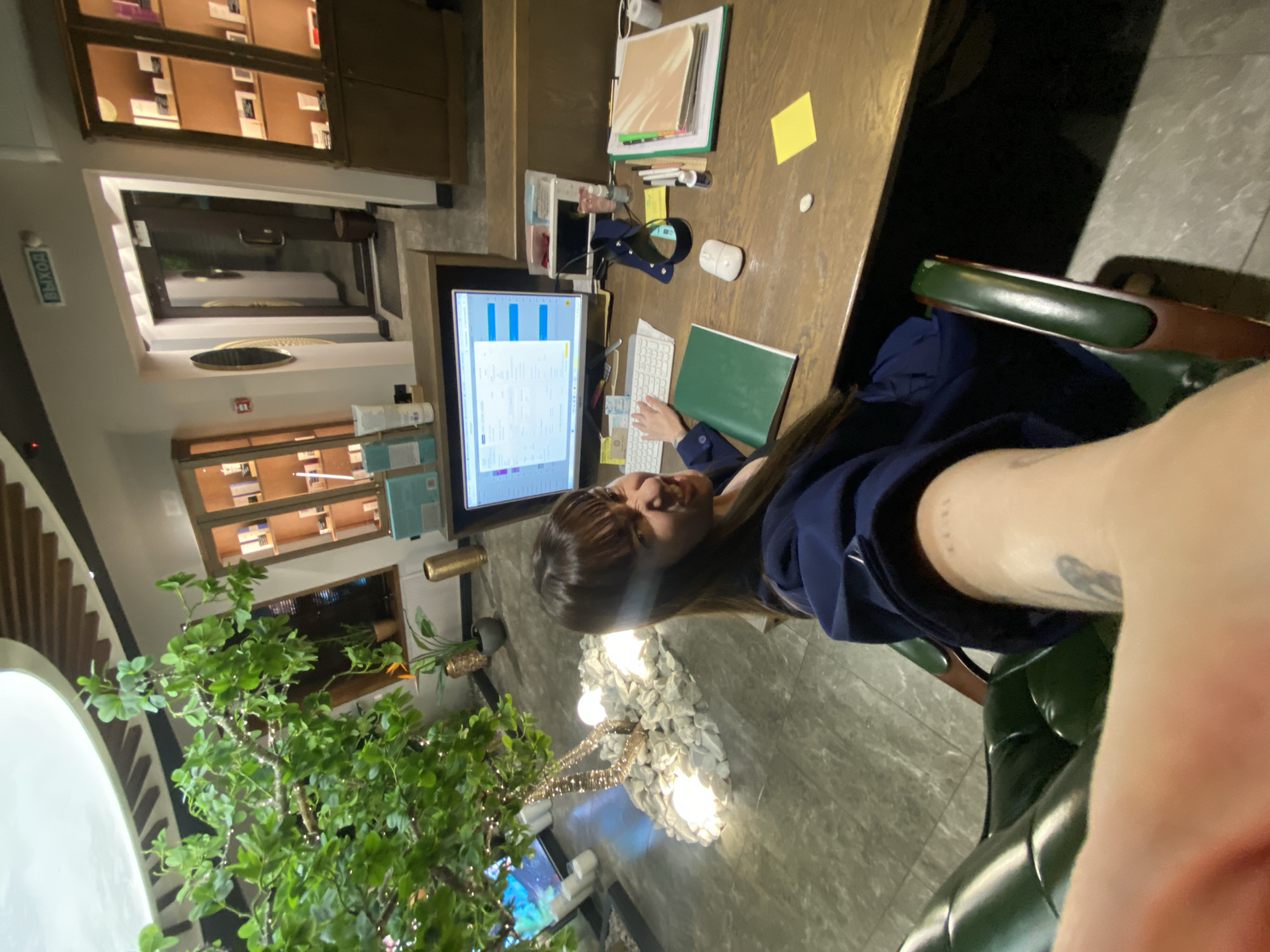 The image captures a woman seated at a desk in what appears to be a public space in Votkinsk, Russia. The desk, constructed of wood, is cluttered with items including a computer monitor displaying charts and graphs, a keyboard, a notebook, and office supplies. The woman, wearing a blue jacket, is focused on the desk, likely working. In the background, shelves display various objects, and a plant adds greenery to the space. The environment suggests a co-working area or a library within the city. The presence of an exit sign suggests an entrance/exit point of the building.