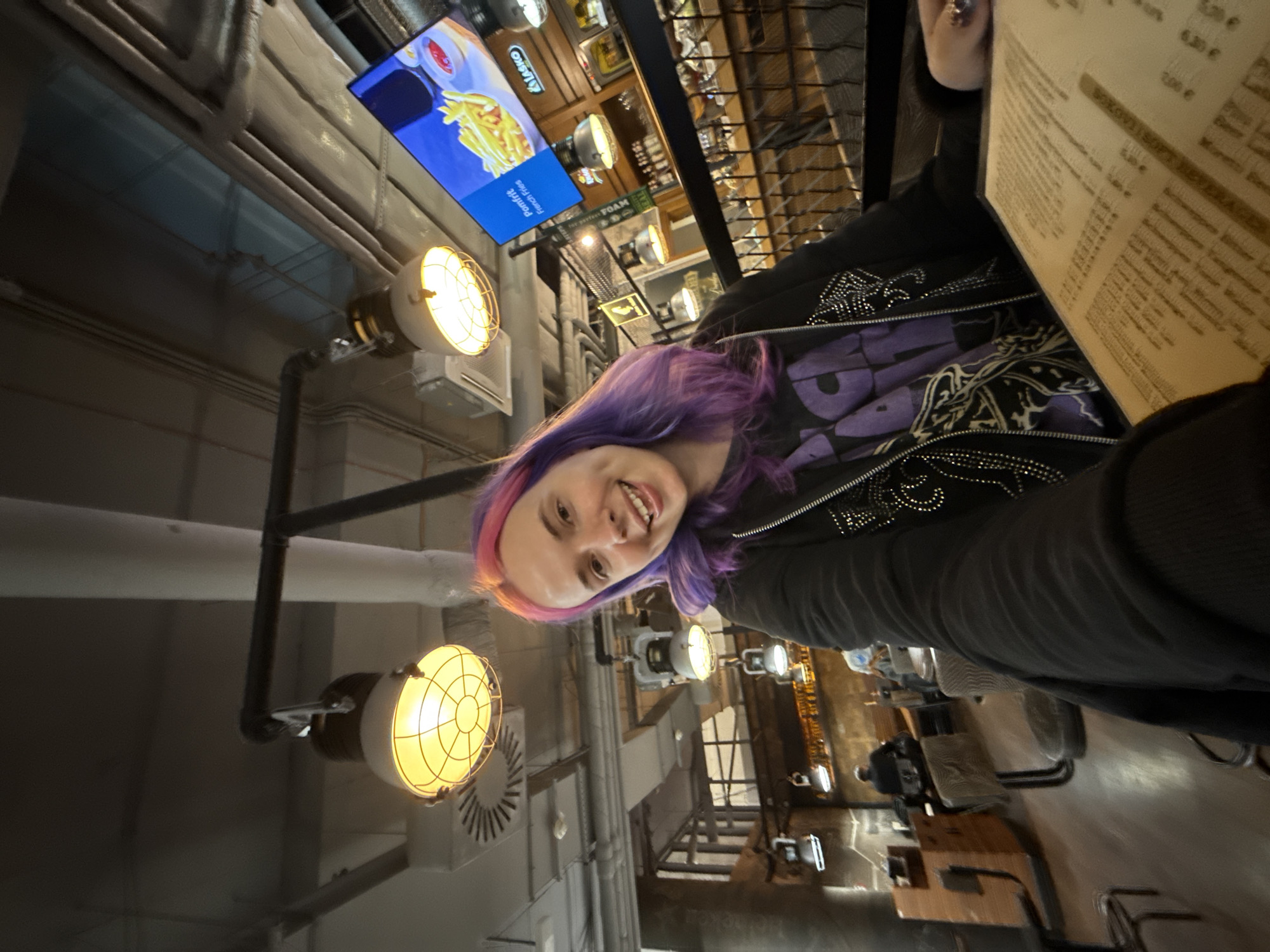 Here is a description of the image:
The image shows a person with purple and pink hair sitting at a restaurant or cafe. The person is looking at the camera and smiling. In front of them is a menu, and there are tables and chairs visible in the background. There are also several industrial-style lights on the ceiling. There is a sign above that says "Pomfrit French Fries" and "LASKO." The image appears to be taken indoors in the city of Velika Gorica, Croatia.